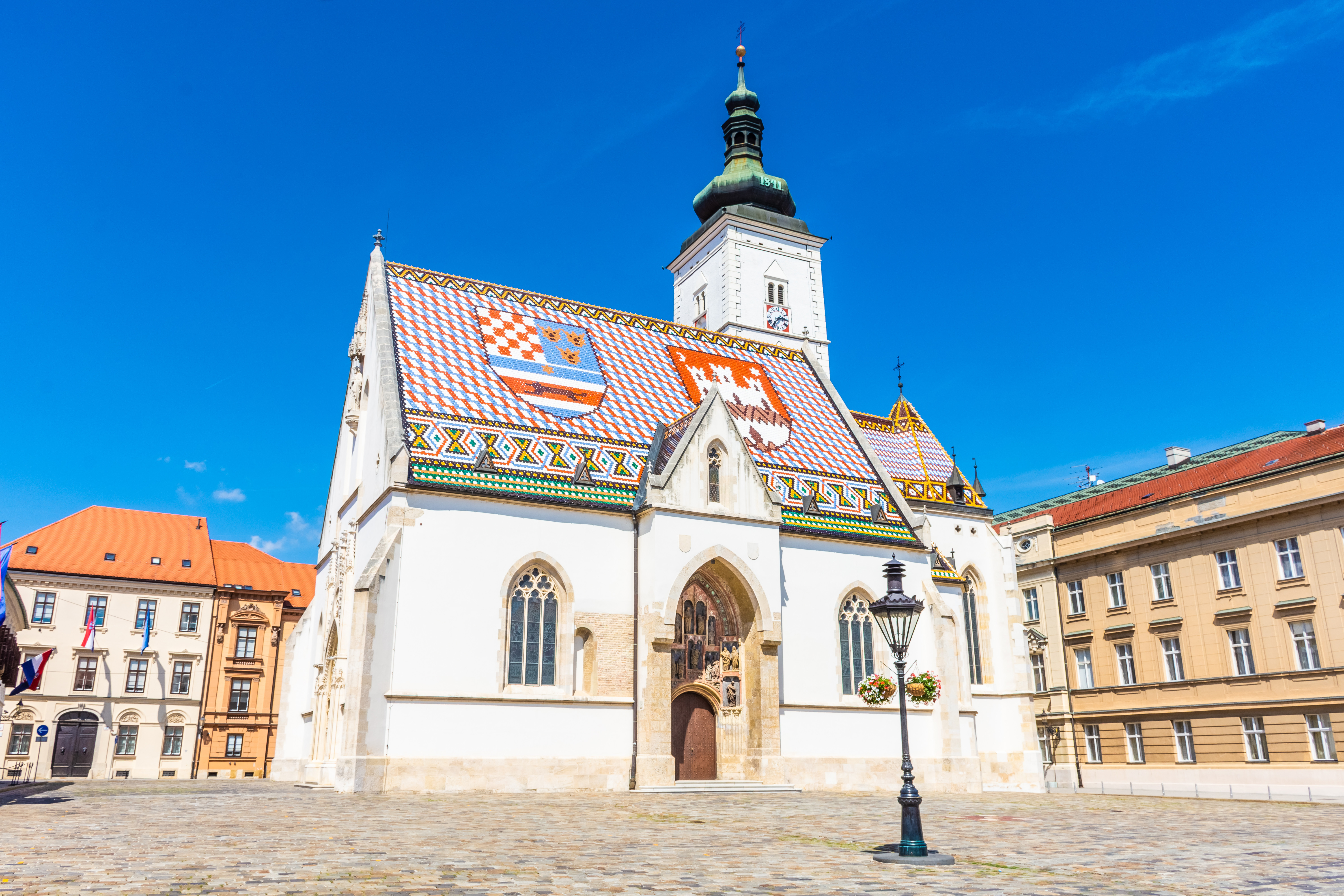St. Markus Kirke i Zagreb med farverigt, mønstret tag under en klar blå himmel, omgivet af historiske bygninger