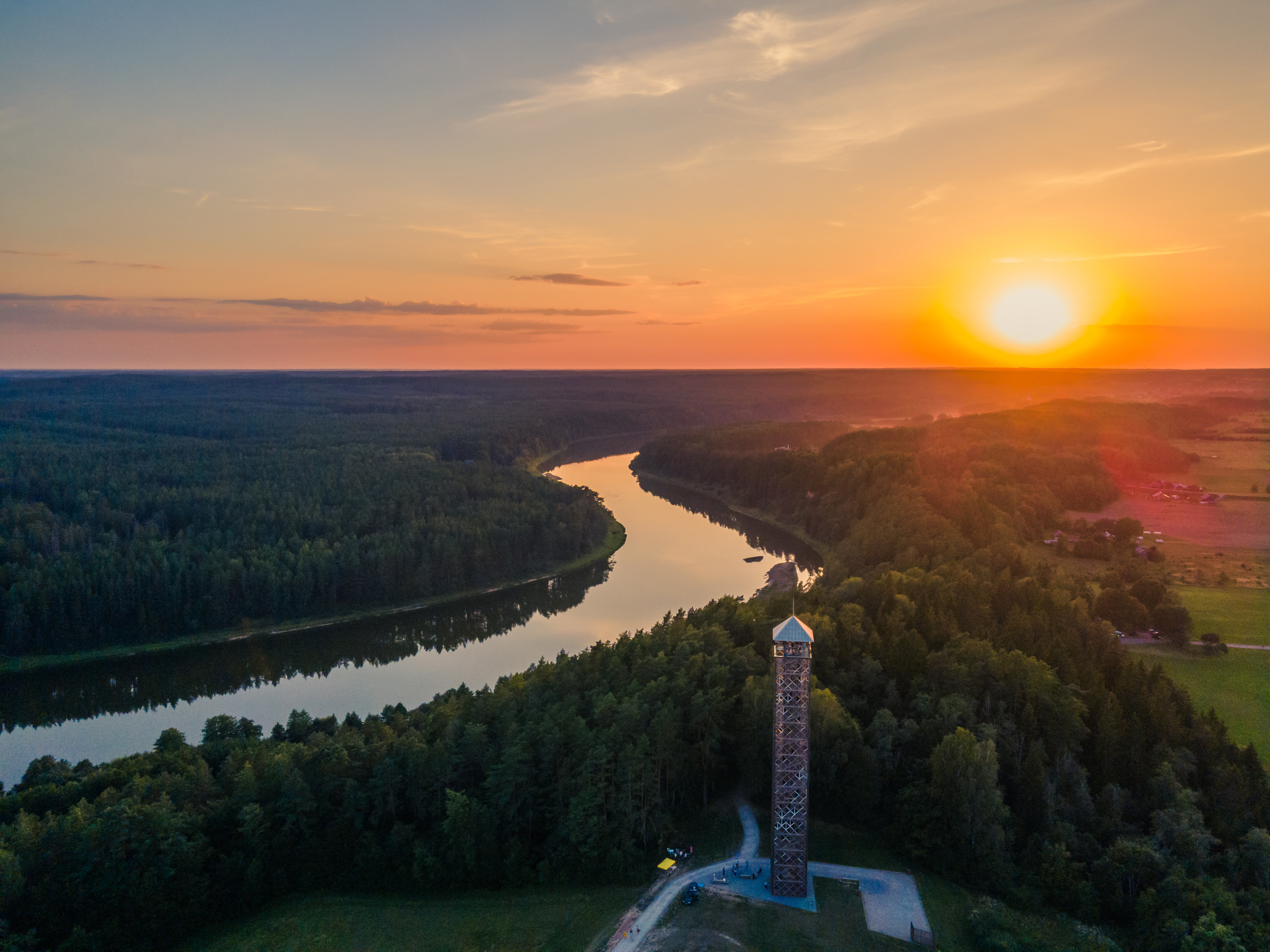 Solnedgang over en naturskøn flod med frodige skove og et højt udsigtstårn, der tilbyder panoramiske udsigter over det livlige landskab