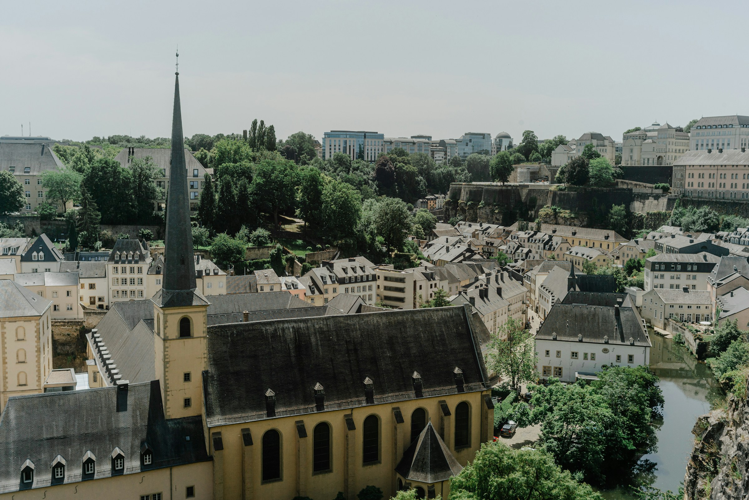 Scenic view of Luxembourg City's historic Grund district with traditional architecture, lush greenery, and a prominent church spire under a clear sky