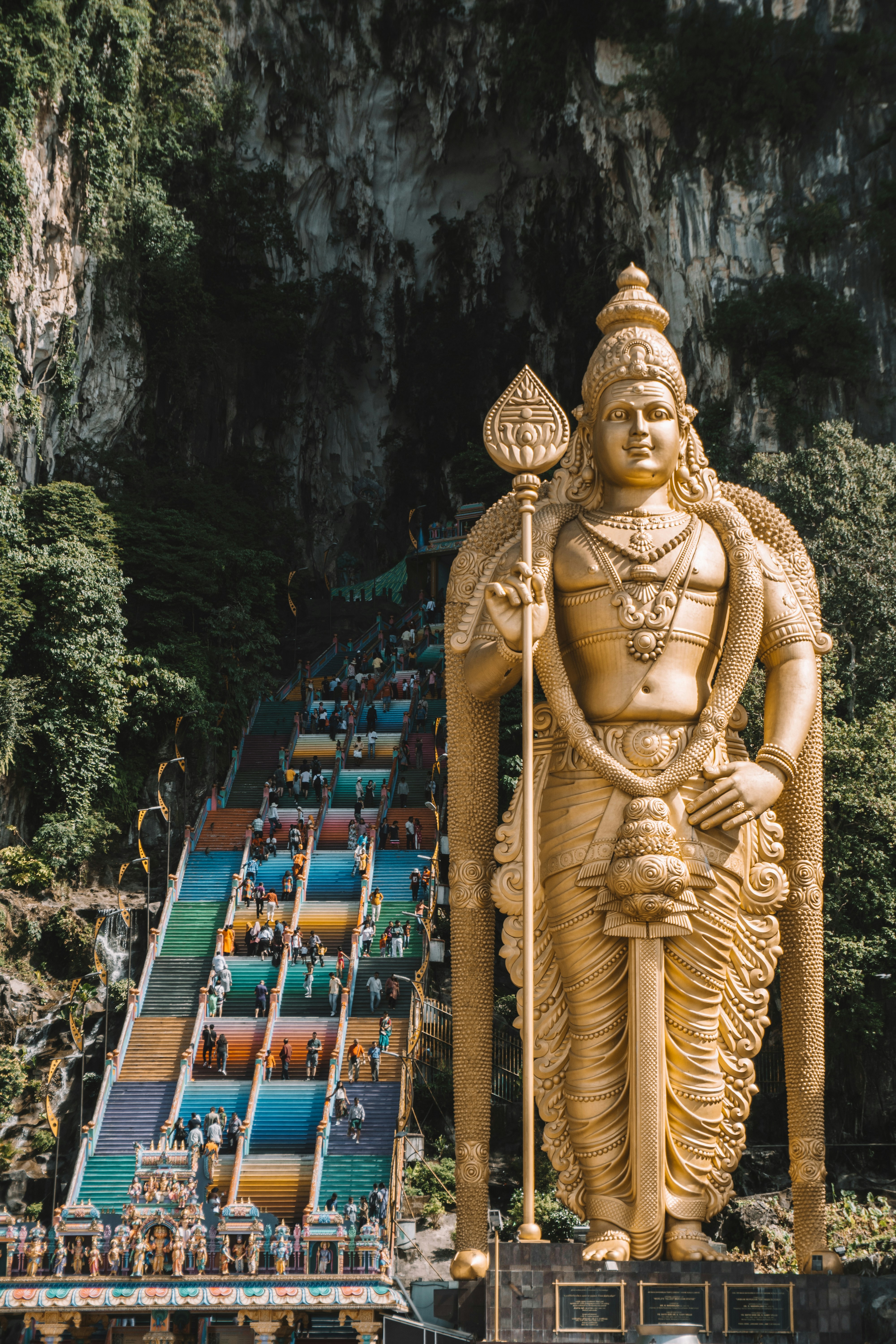 Gylden statue af Lord Murugan ved indgangen til Batu Caves, Malaysia, med en farverig trappe, der fører til grottetemplet, omgivet af frodige grønne klipper