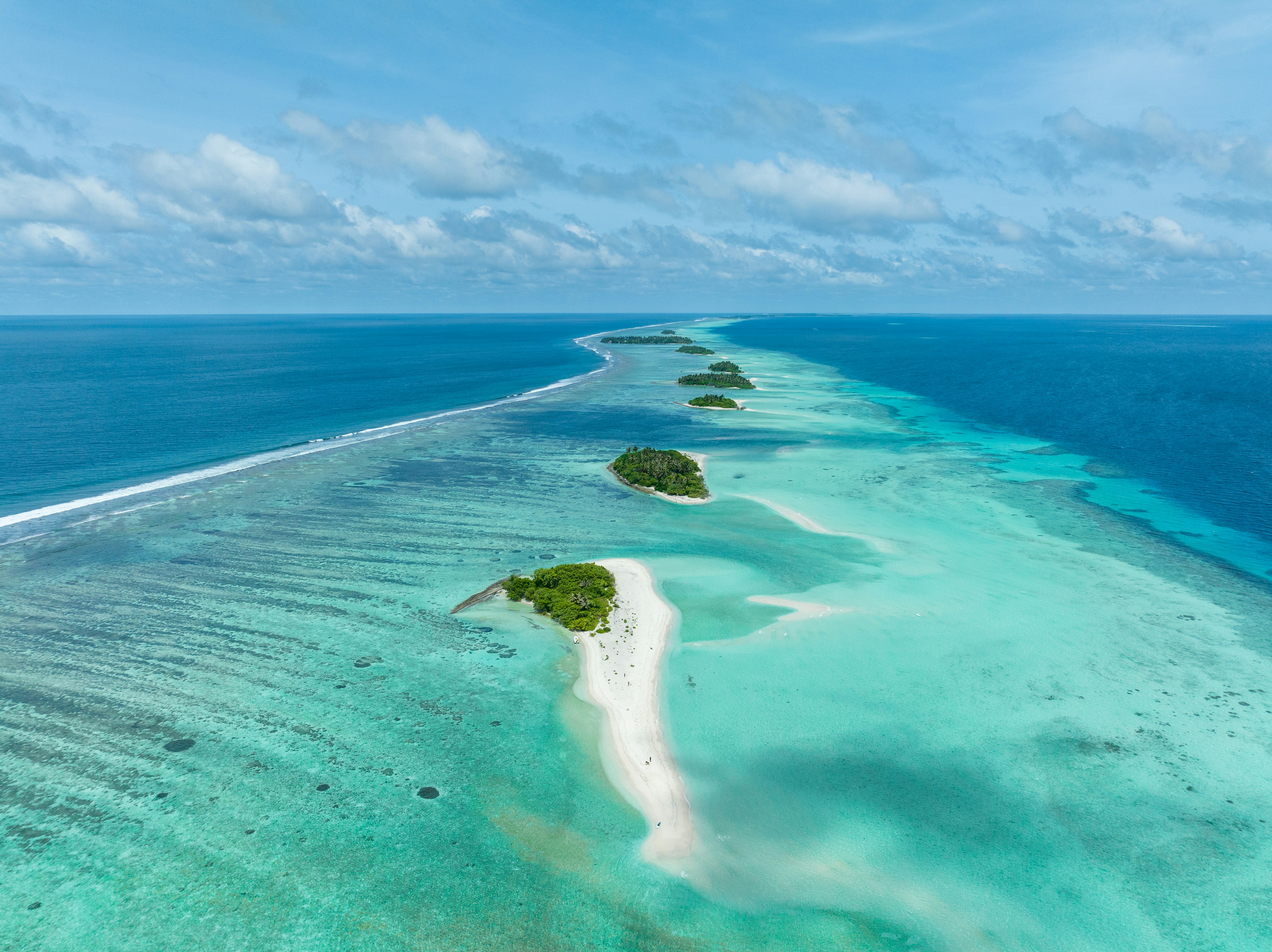 Luftfoto af et tropisk atoll med frodige grønne øer og hvide sandstrande omgivet af turkisblåt hav under en blå himmel