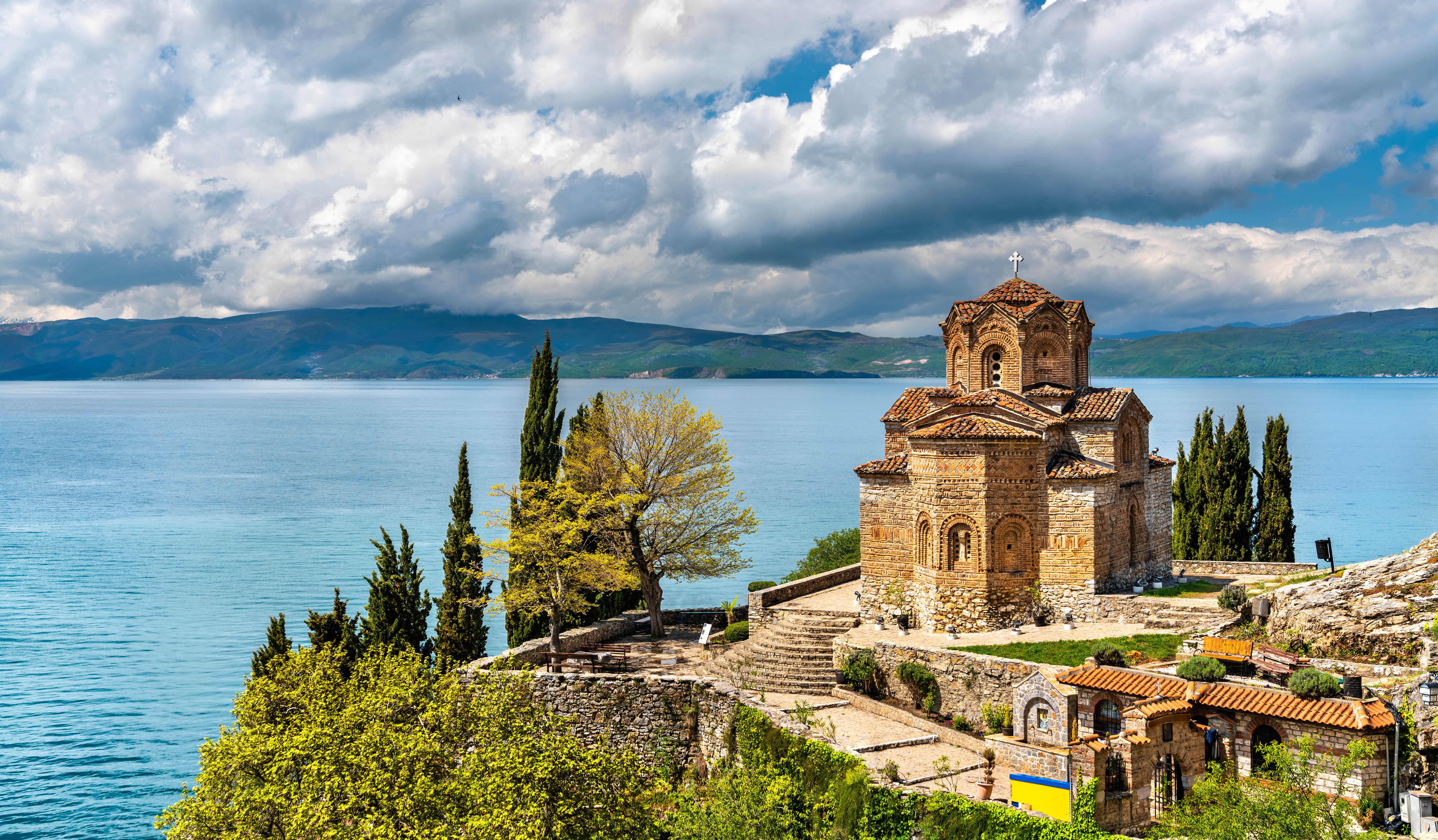Rejser til Nordmakedonien - Gammel kirke beliggende ved bredden af en sø med bjerge i baggrunden og en skyet himmel.