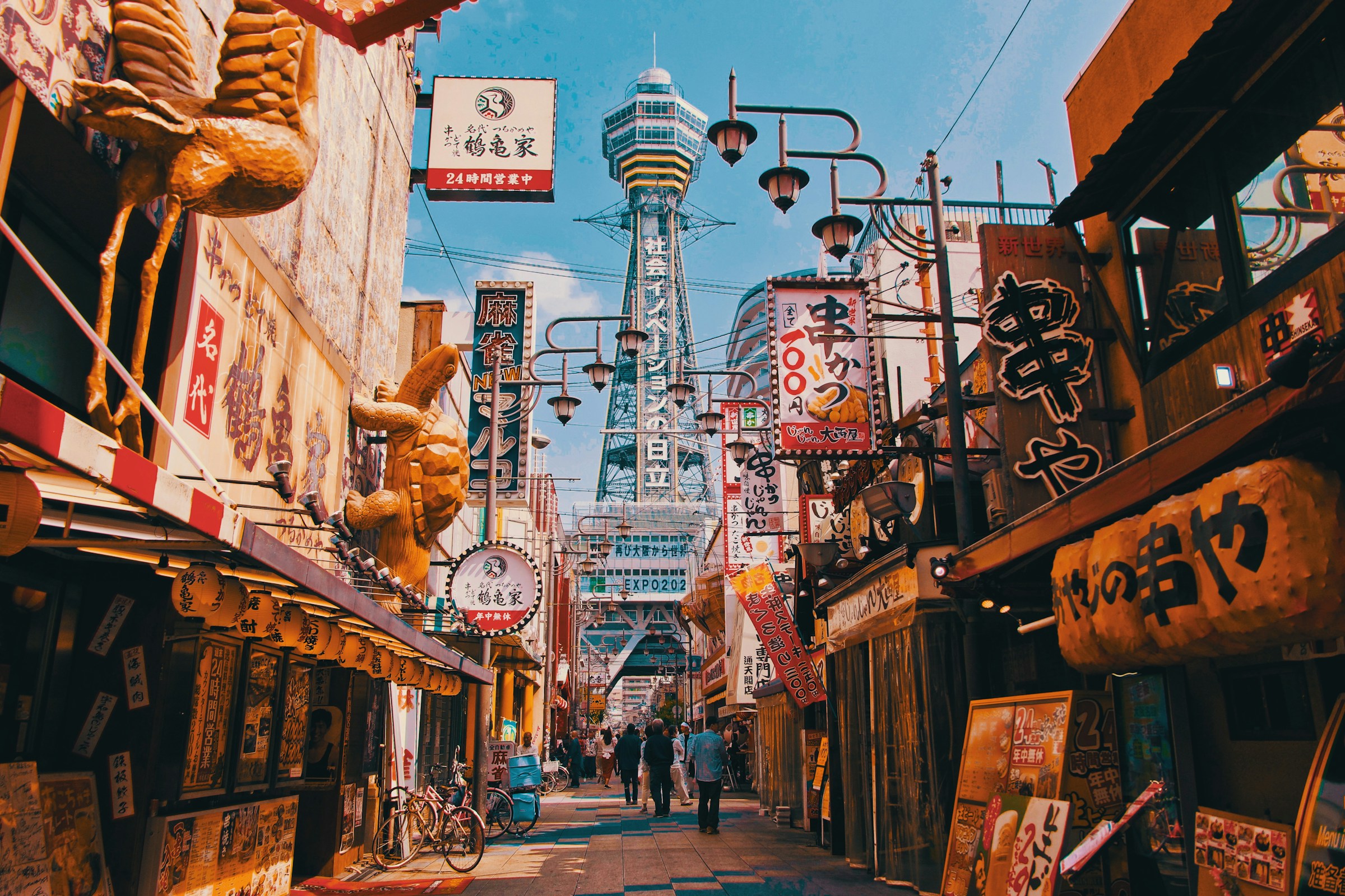 Travl gade i Osaka, Japan, med restauranter, skilte og udsigt til Tsutenkaku-tårnet under klar blå himmel.