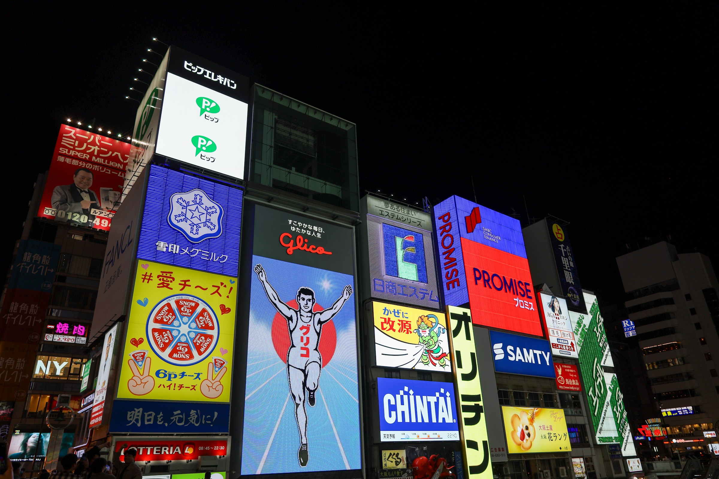 Neonskilte og reklamer på bygninger i Dotonbori, Osaka, Japan, med Glica som ophænger med skilt i midten, natteudsigt.