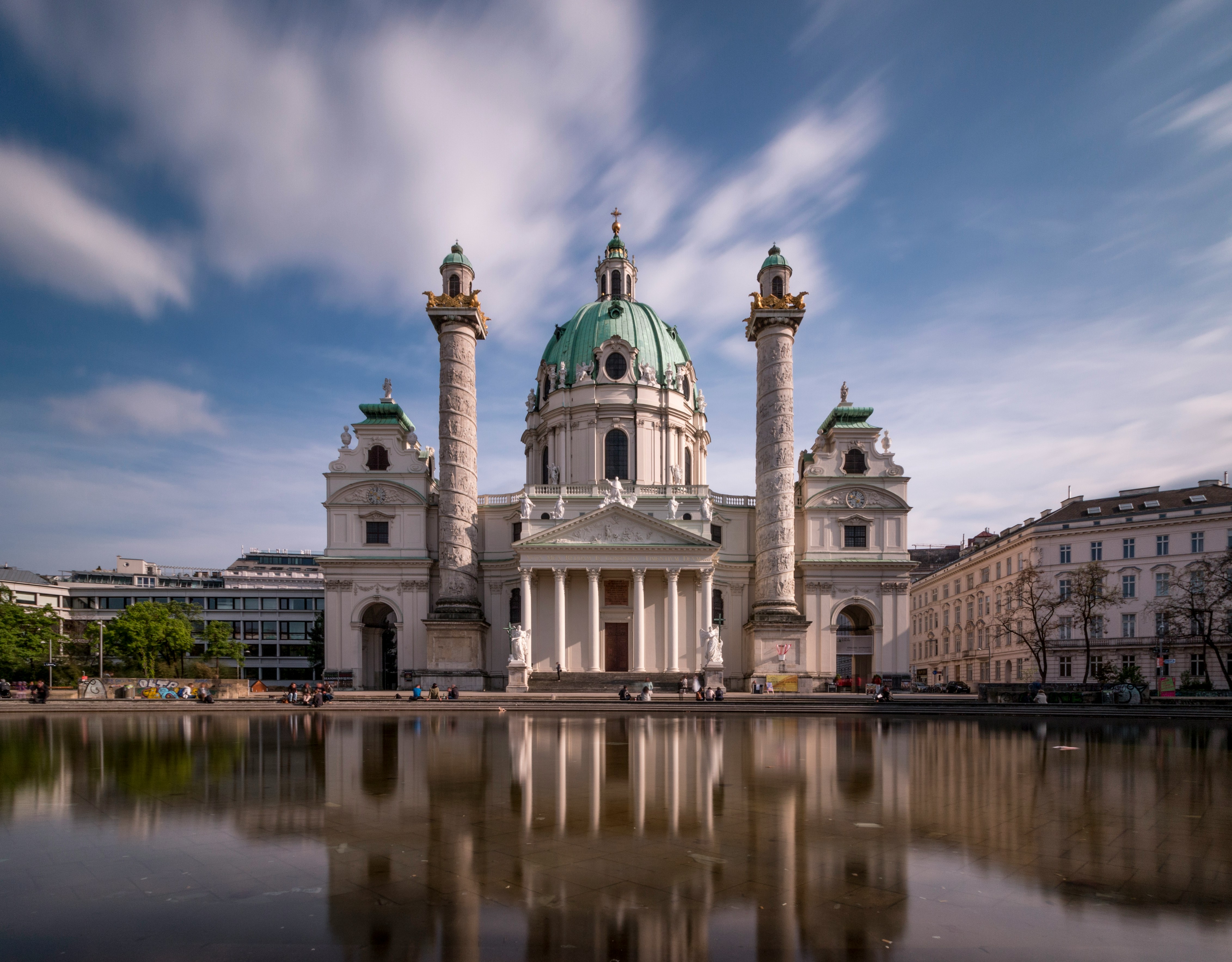 St. Charles Kirke i Wien, Østrig, med sin barokarkitektur og karakteristiske grønne kuppel, der spejler sig i en rolig pool under en blå himmel