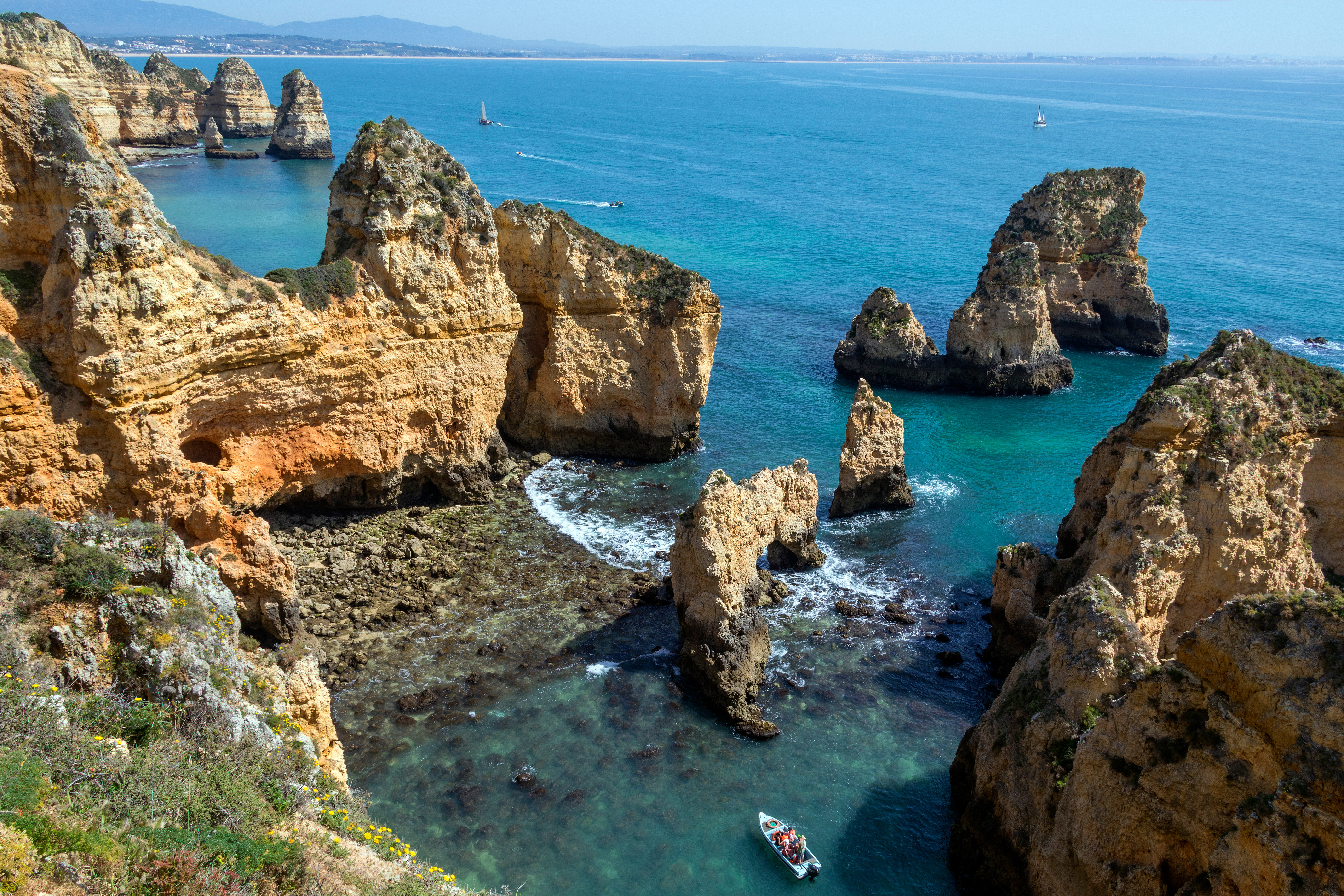 Scenisk udsigt over Ponta da Piedade-klipperne i Lagos, Portugal, med imponerende kalkstensformationer omgivet af turkisfarvet vand, hvor både og fjerne sejlskibe tilføjer charme til kystlandskabet
