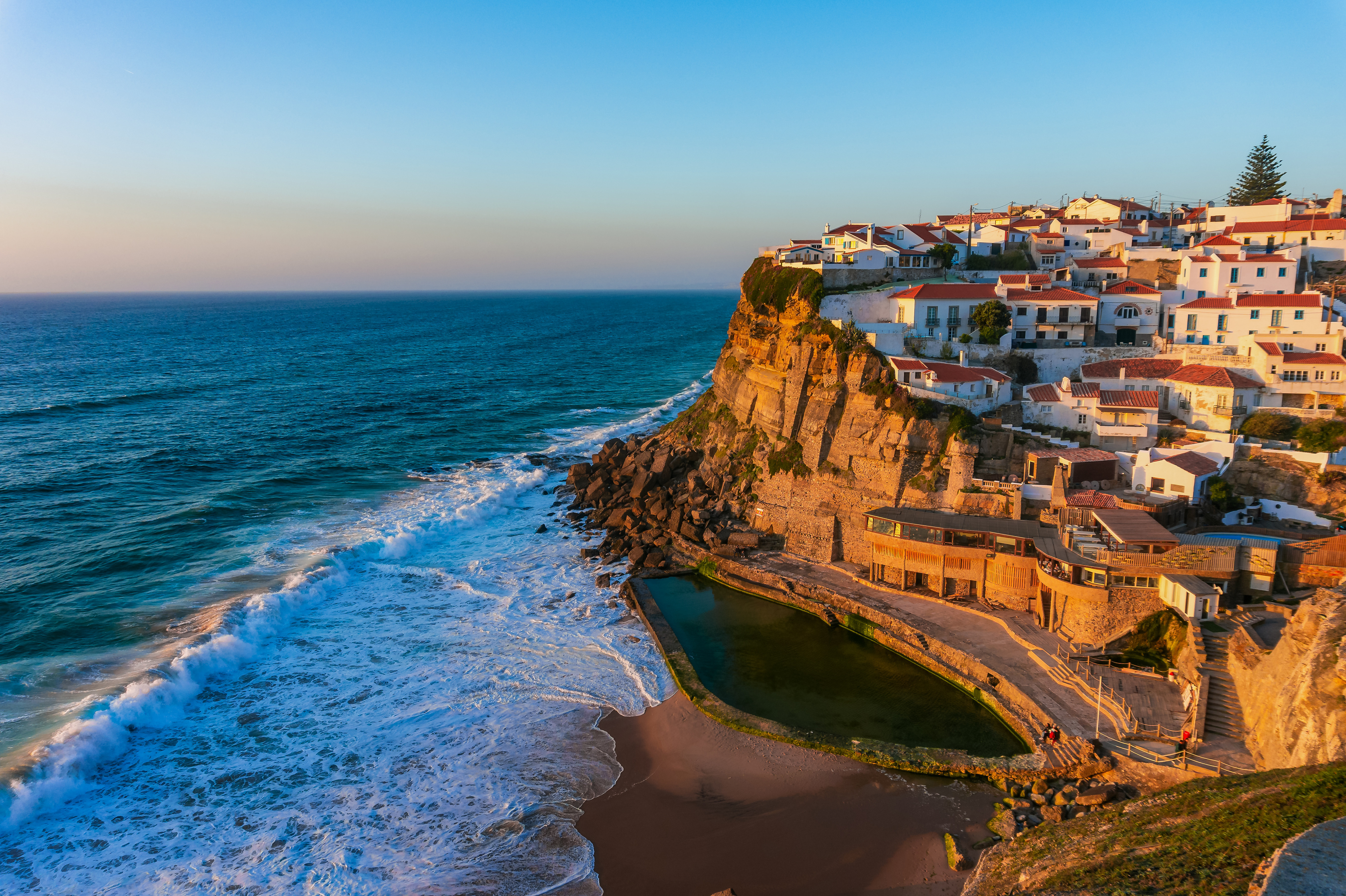 Scenisk udsigt over Azenhas do Mar, Portugal, der viser hvide huse placeret på en klippe over Atlanterhavet ved solnedgang, med bølger der brydes nedenfor og en naturlig swimmingpool i forgrunden