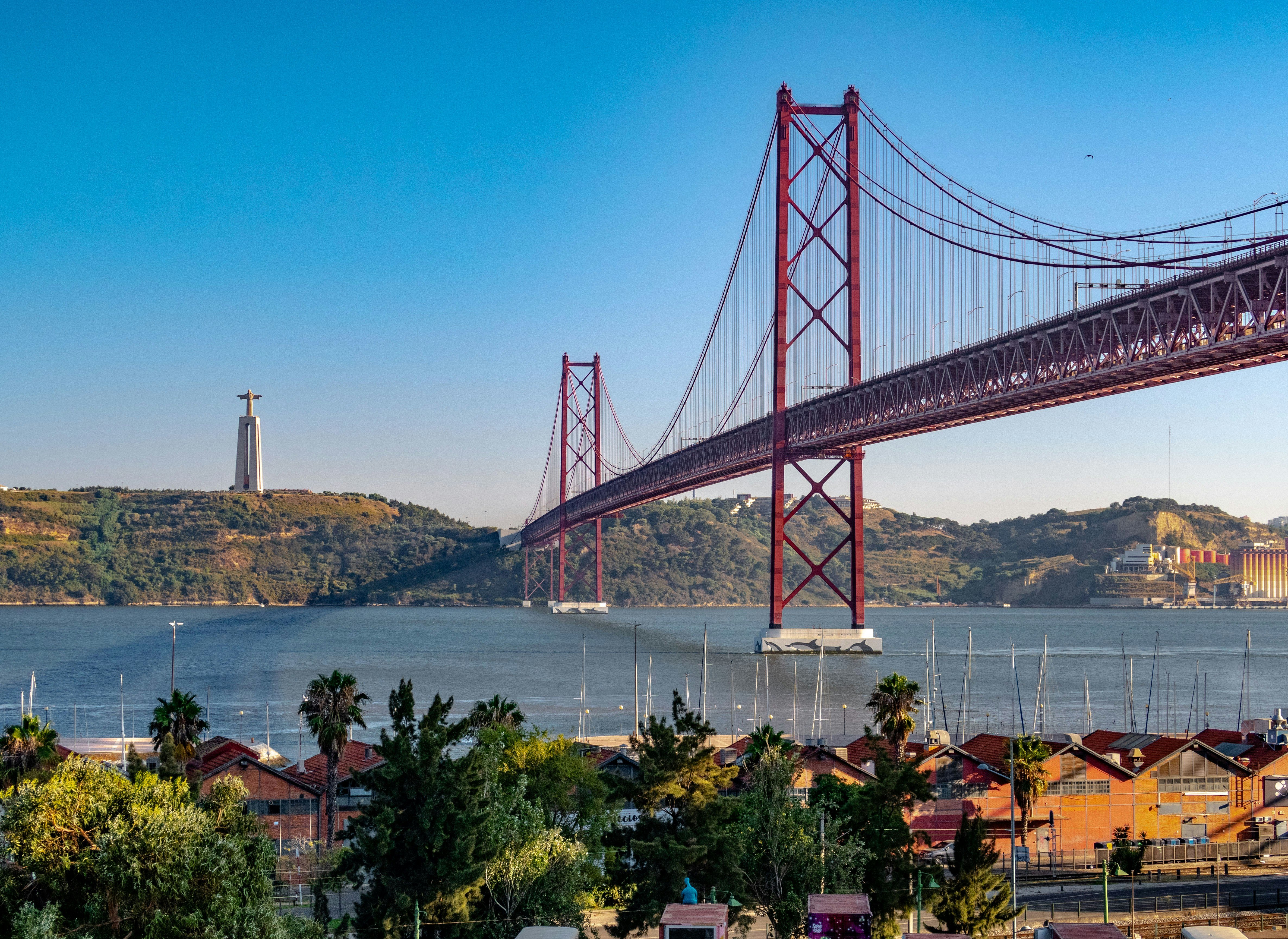 25 de Abril-broen, der spænder over Tejo-floden i Lissabon, Portugal, med Cristo Rei-statuen synlig til venstre under en klar blå himmel
