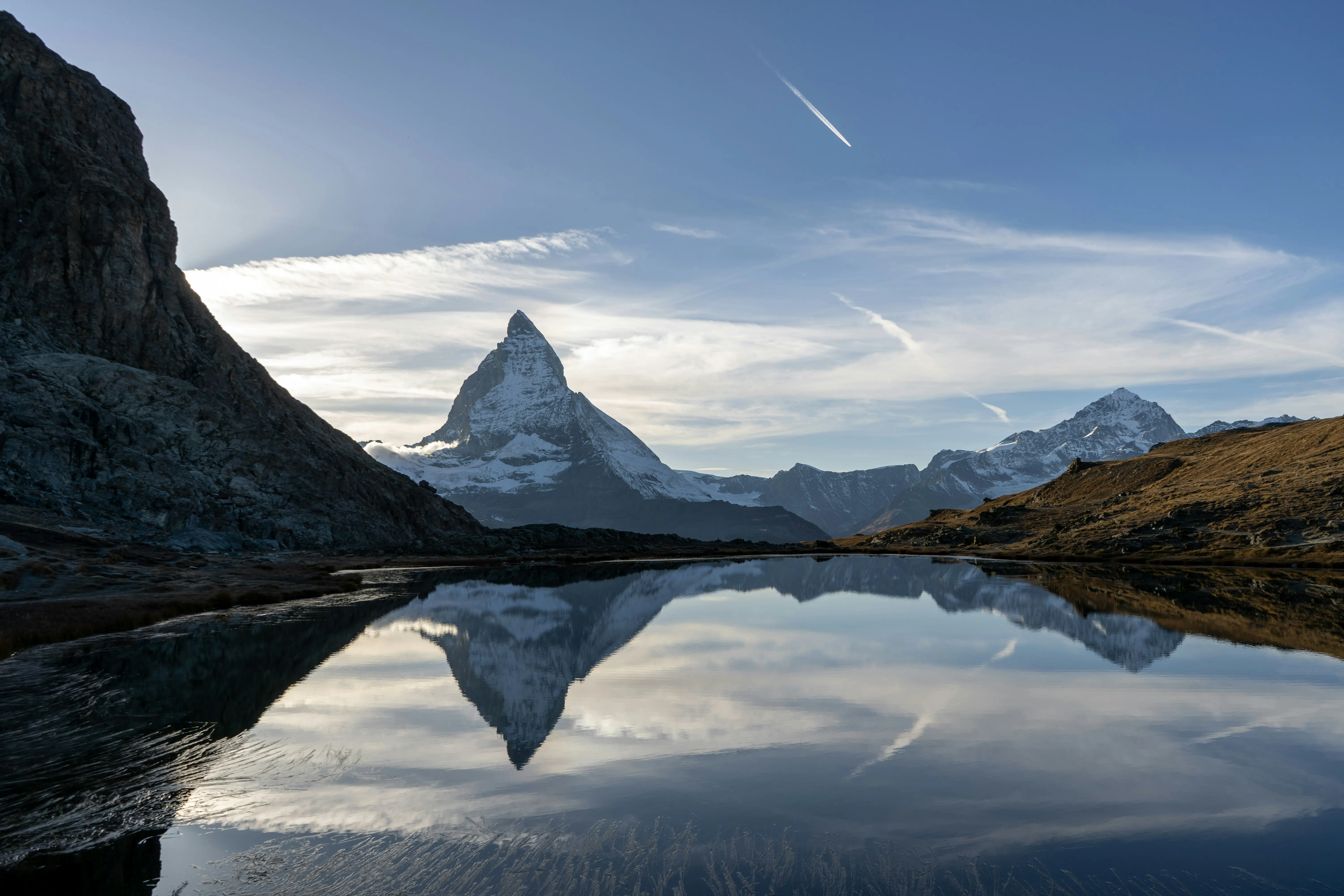 Matterhorn-bjerget med sneklædt top spejlet i en rolig sø under en klar blå himmel med skyer, omgivet af et barsk terræn