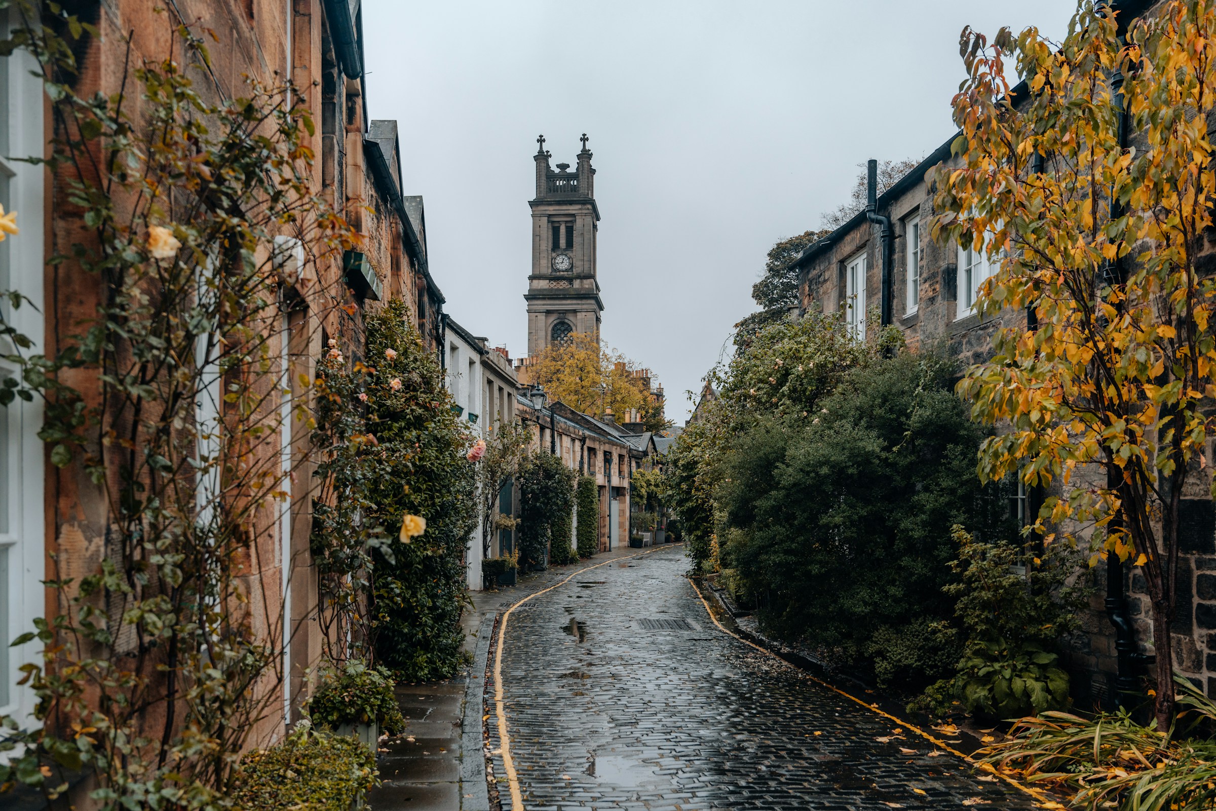 Quaint cobblestone street in Edinburgh with historic buildings and a church tower in the background, surrounded by lush greenery and autumn foliage.