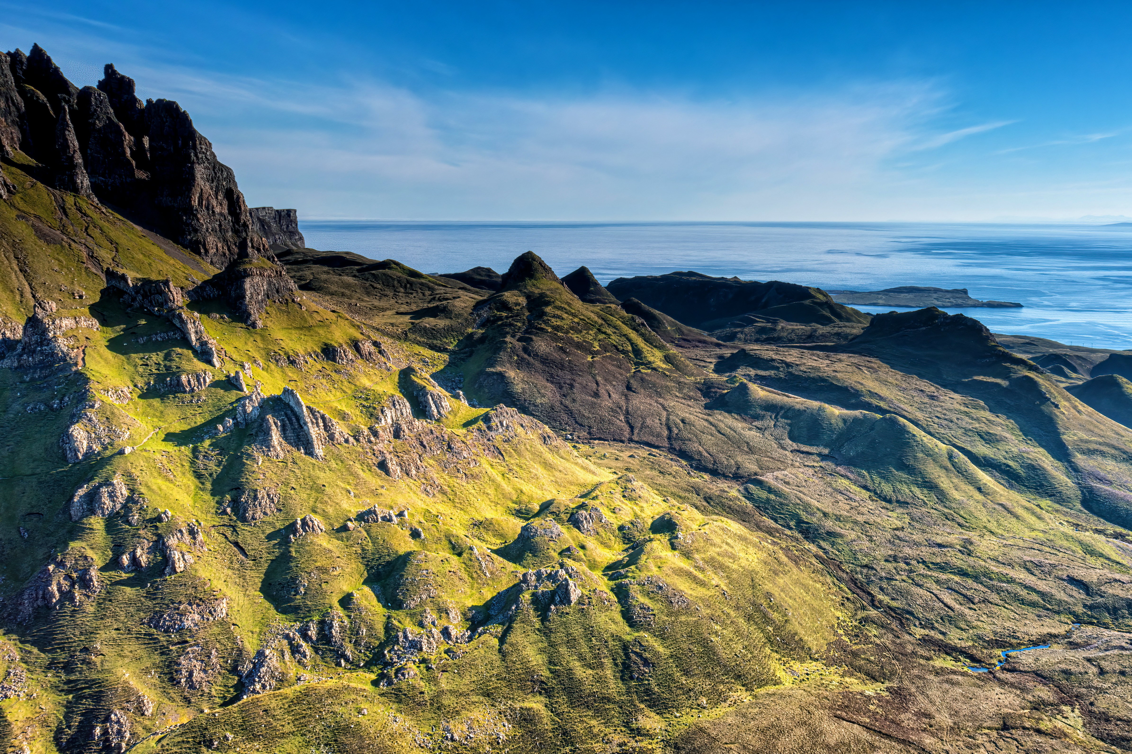 Malende luftfoto af Quiraing-skredet på Isle of Skye i Skotland, med bølgende bakker dækket af livligt grønt græs under en klar blå himmel og udsigt over havet
