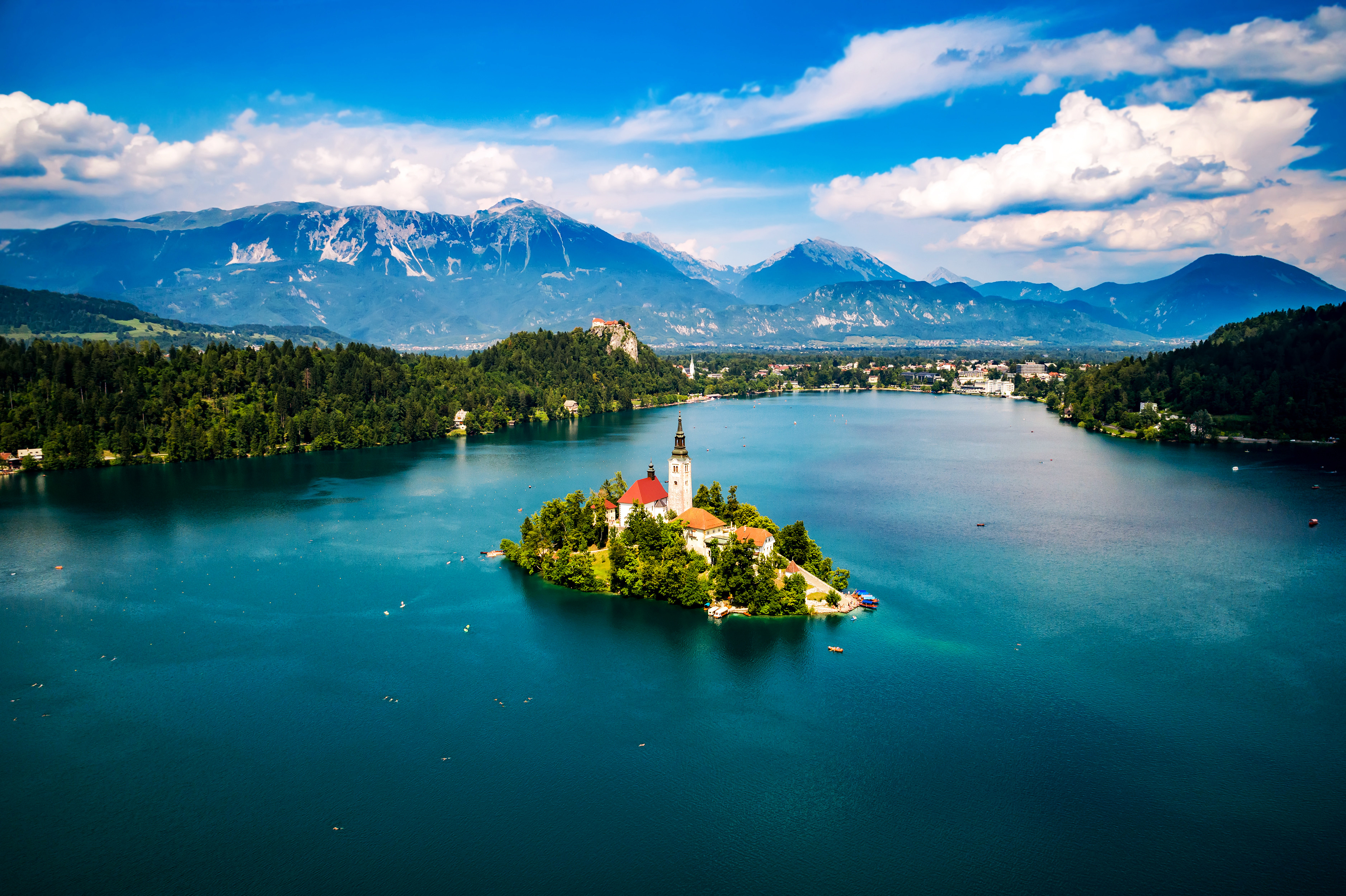 Skøn udsigt over Lake Bled med en kirkekirke på en ø, omgivet af klart blåt vand og frodig grøn natur, med Julian Alperne i baggrunden under en delvist overskyet himmel