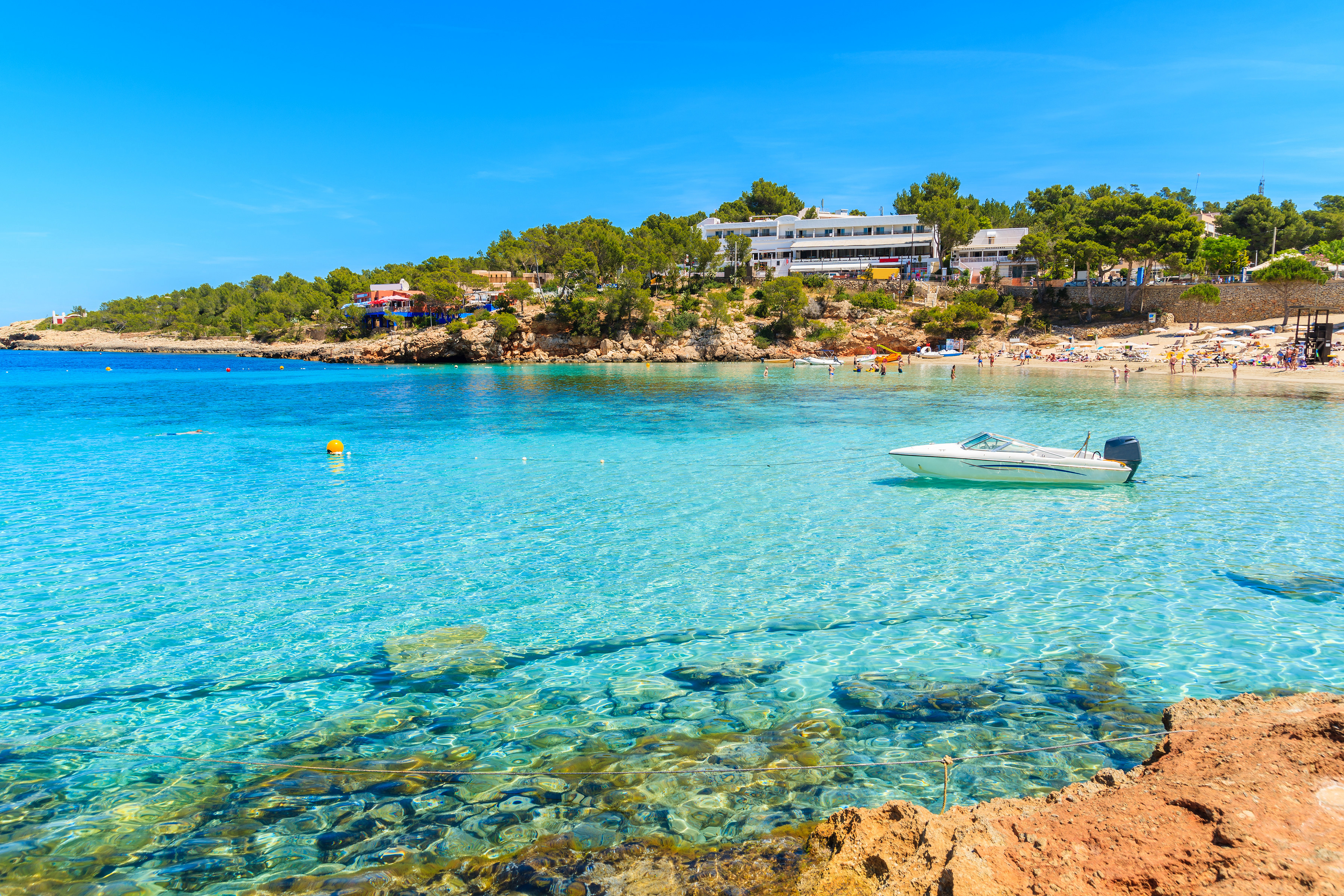 A scenic view of a clear turquoise bay with a small motorboat and a rocky shoreline, surrounded by lush greenery and a sandy beach in the background, under a bright blue sky