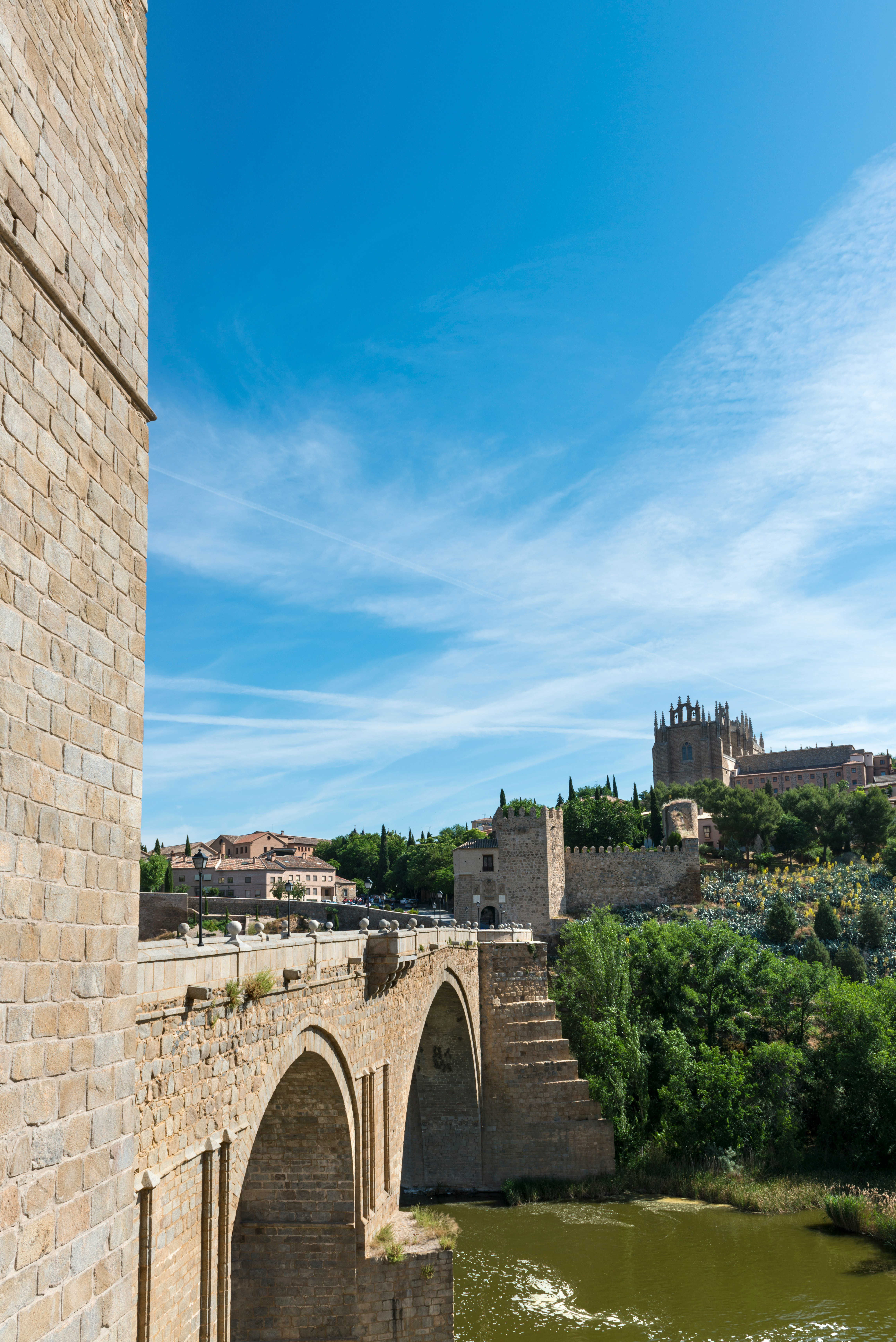 Historisk stenbro over flod i Toledo, Spanien, med middelalderlig arkitektur og frodig grøn vegetation under en klar blå himmel