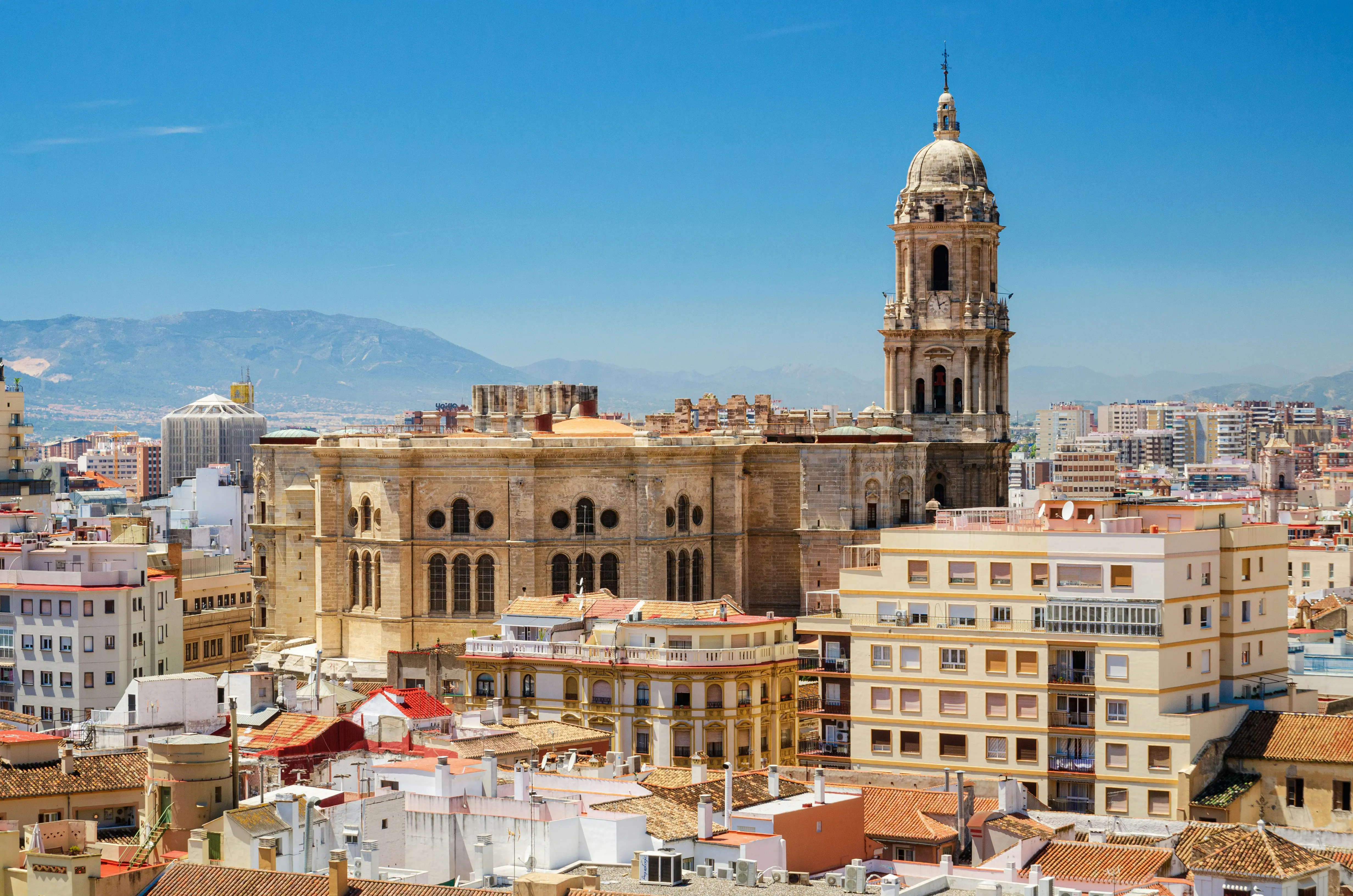 Udsigt over Malaga Katedrals arkitektur i bybilledet i Malaga, Spanien, med omkringliggende bygninger og et bjergrigt baggrund under en klar blå himmel
