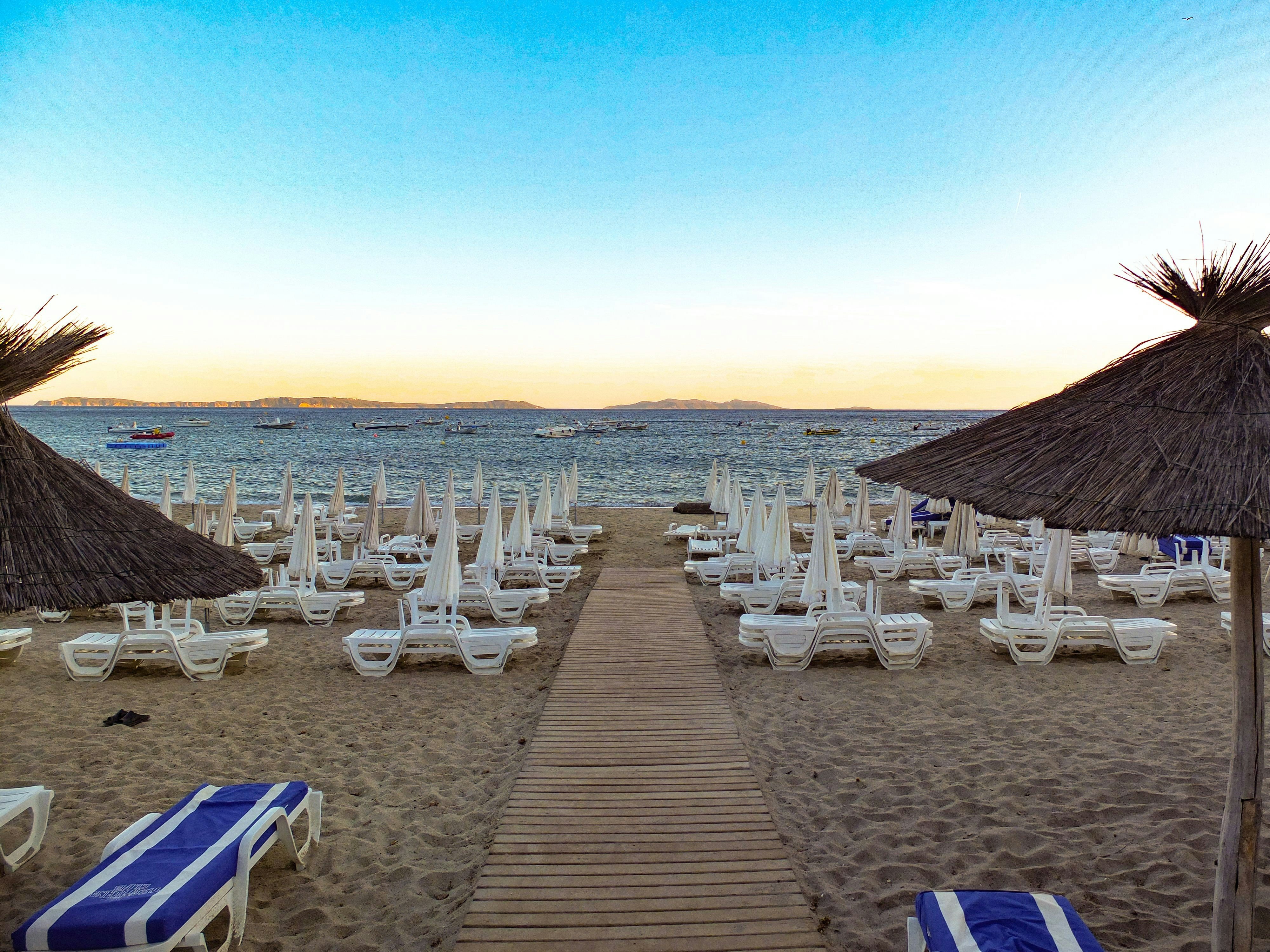 Sandy beach with rows of empty lounge chairs and umbrellas, overlooking a calm sea with several boats near the horizon under a clear blue sky