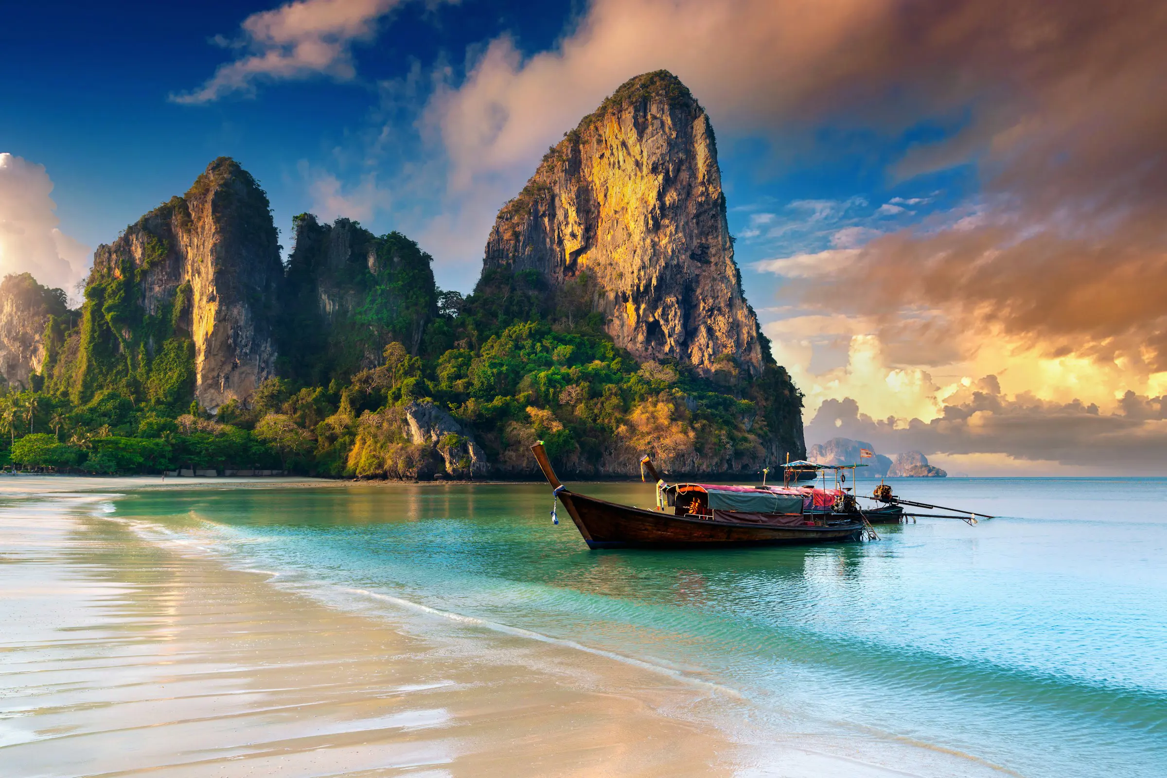 Tidlig skumring ved en strand, der møder blåt hav og en longtailbåd med bjerge i baggrunden i Thailand