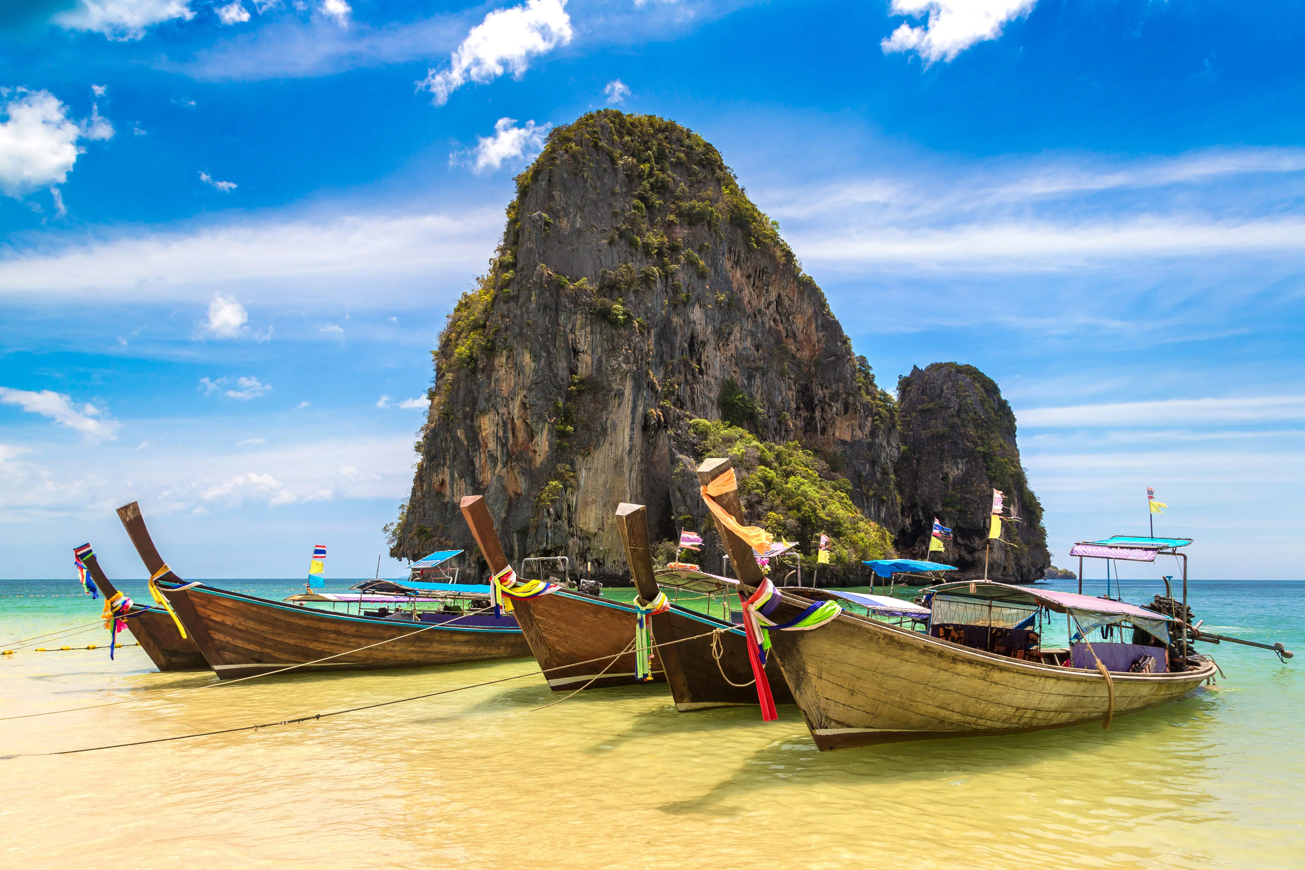 Rejs til Thailand - Longtail-både på en tropisk strand med klippeformationer i baggrunden, klar blå himmel og turkisblåt hav i Thailand.