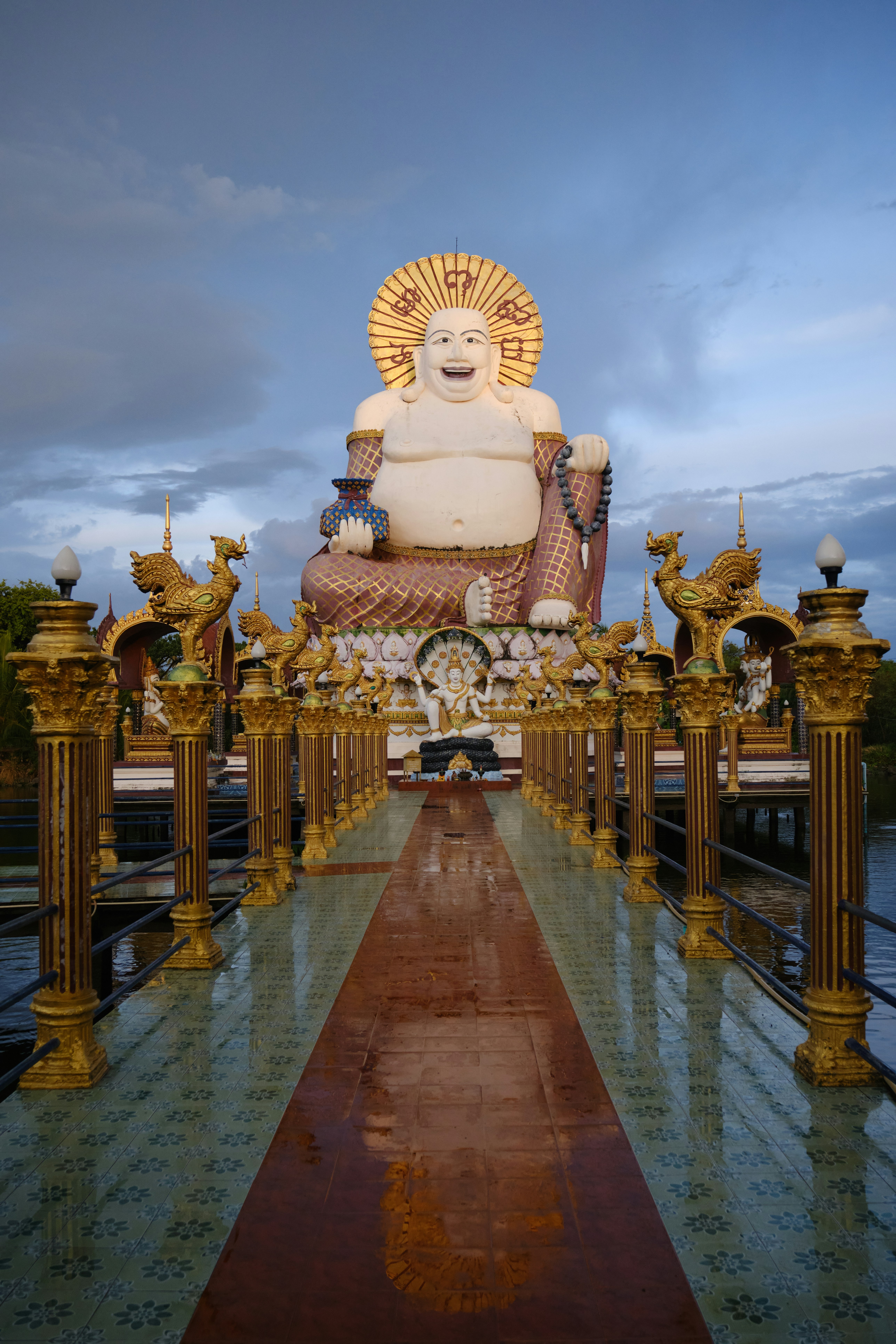 Stor hvid Buddha-statue med gyldne dekorationer ved Wat Plai Laem-templet på Koh Samui, Thailand, set fra en udsmykket gangbro med pragtfulde søjler under solnedgang