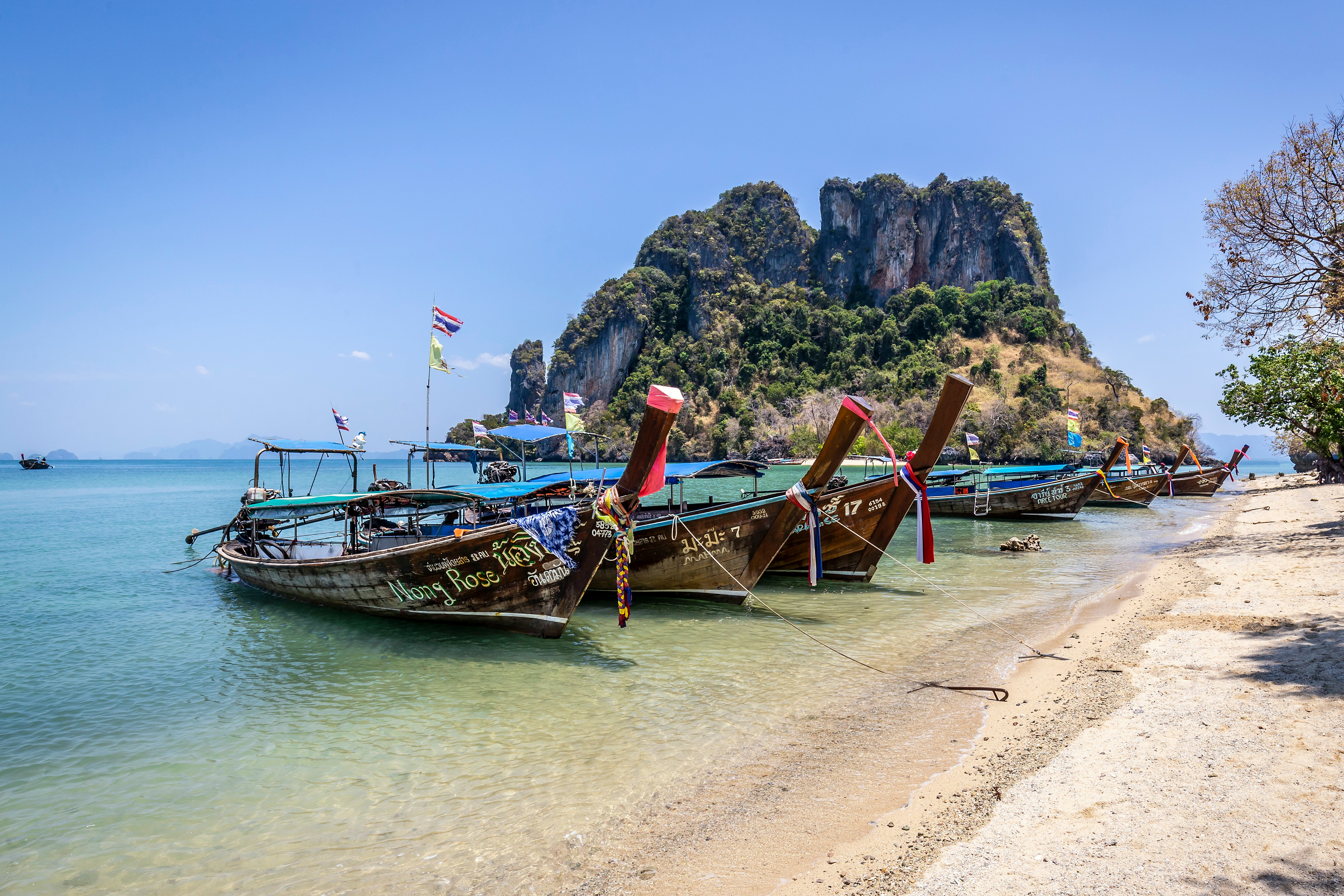 Traditionelle thailandske longtailbåde ved en strand i Krabi, Thailand, med kalkstensklipper i baggrunden under en klar blå himmel