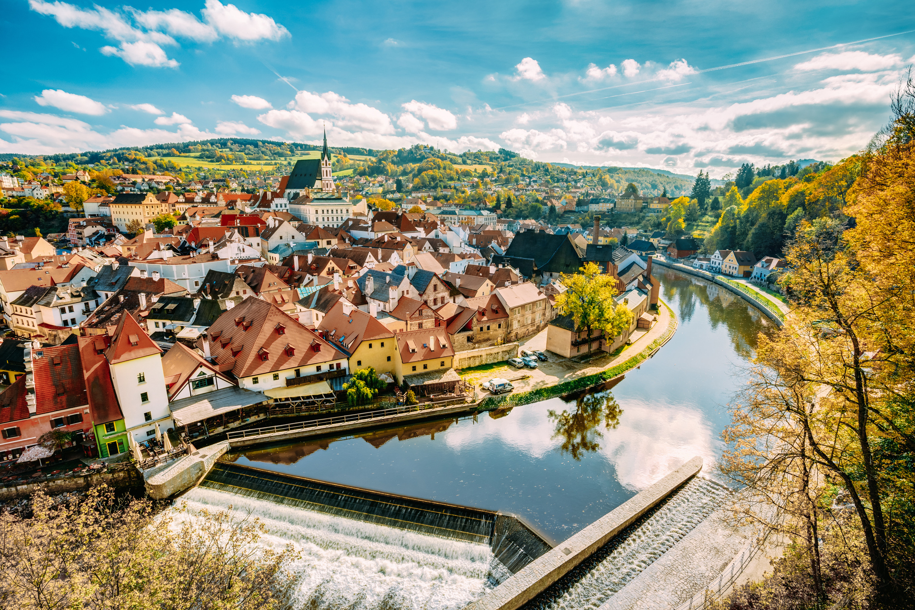 Panoramaudsigt over Český Krumlov, en historisk by i Tjekkiet, med charmerende middelalderarkitektur, floden Vltava og frodigt efterårsløv under en blå himmel