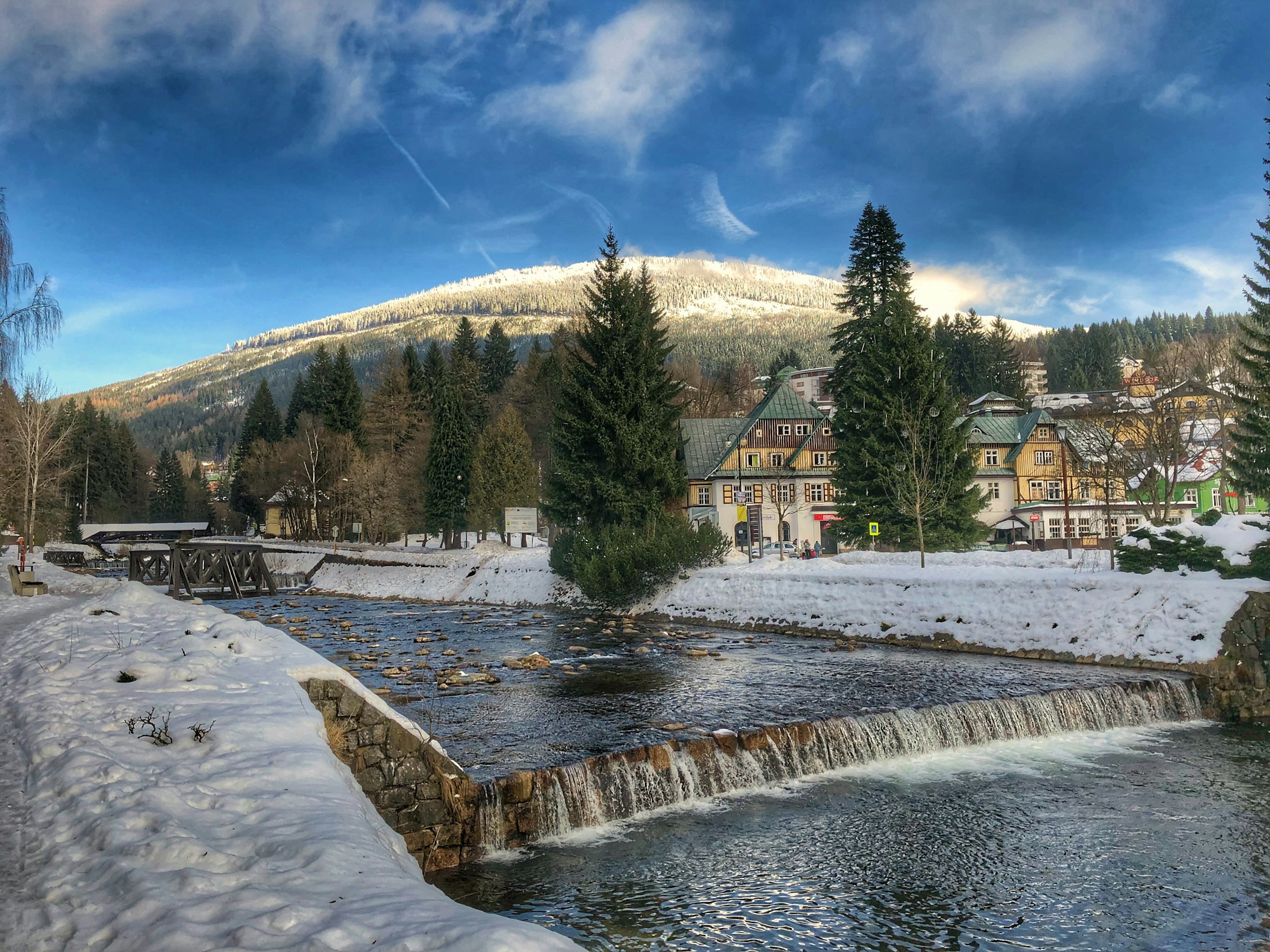 En flod løber gennem en lille snedækket landsby i Spindleruv Mlyn med bjerge og blå himmel i baggrunden