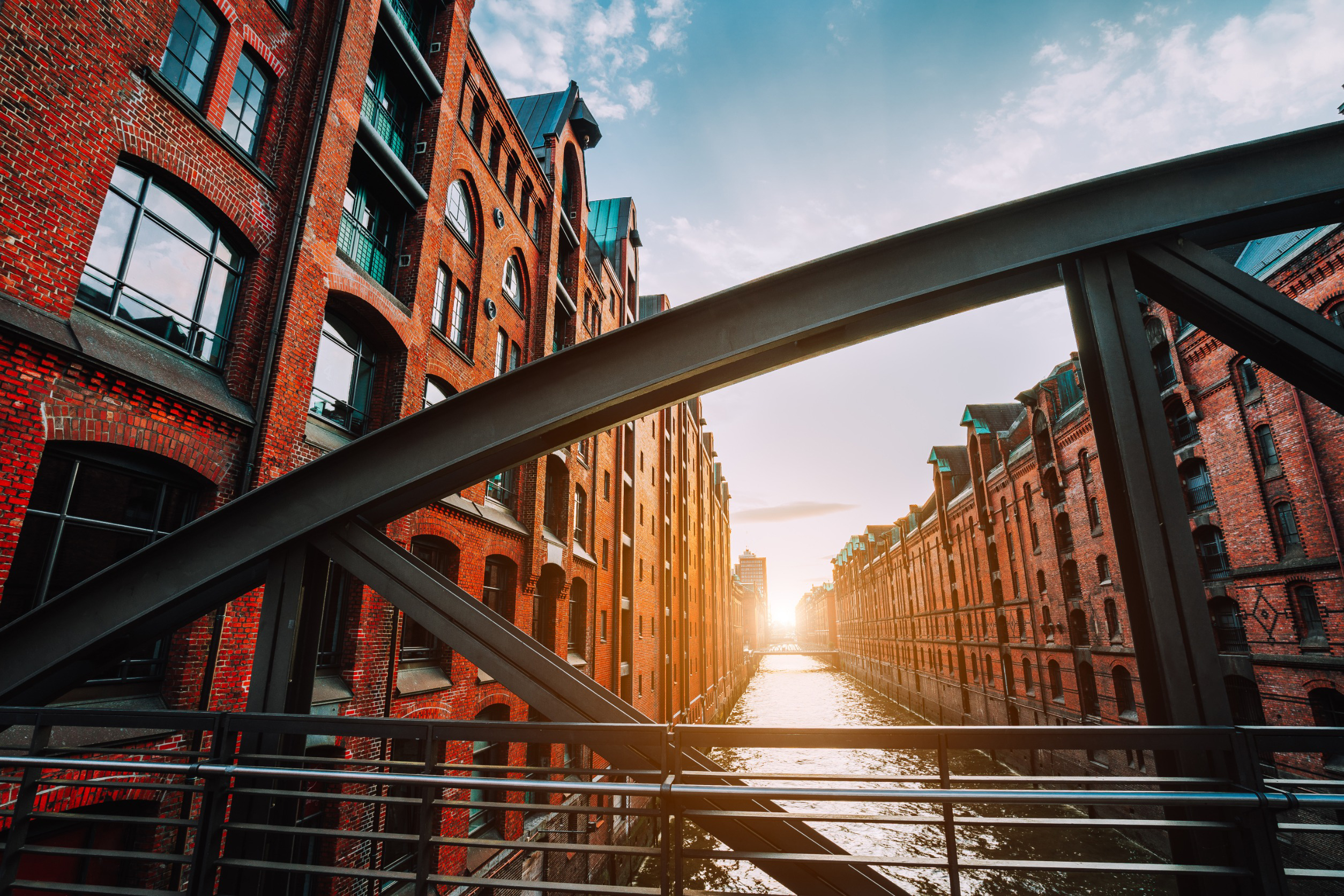 Historic brick warehouse district with canal, viewed from a metal bridge at sunset, featuring warm sunlight and blue sky.