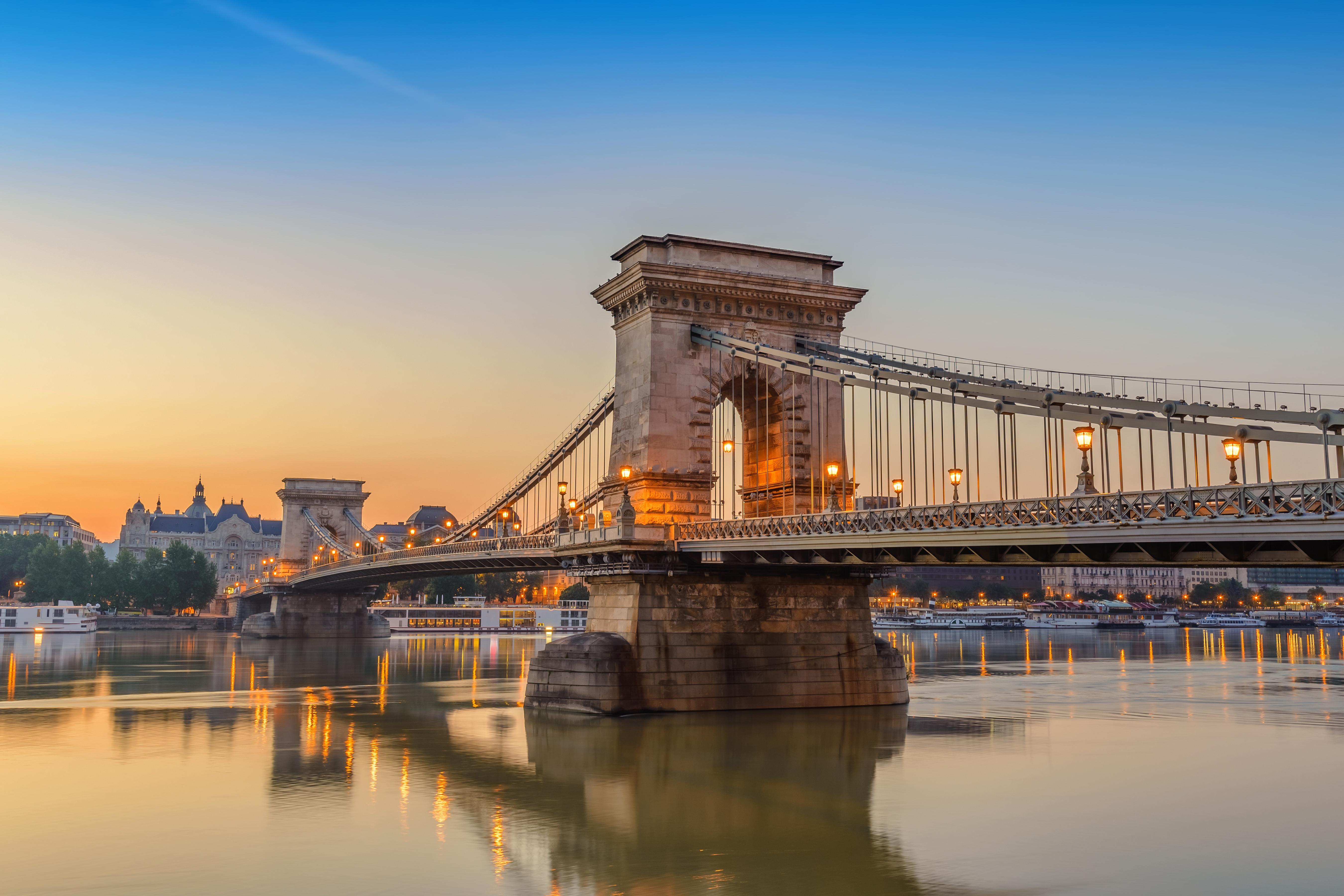 Chain Bridge in Budapest at sunset, reflecting over the Danube River with city buildings in the background