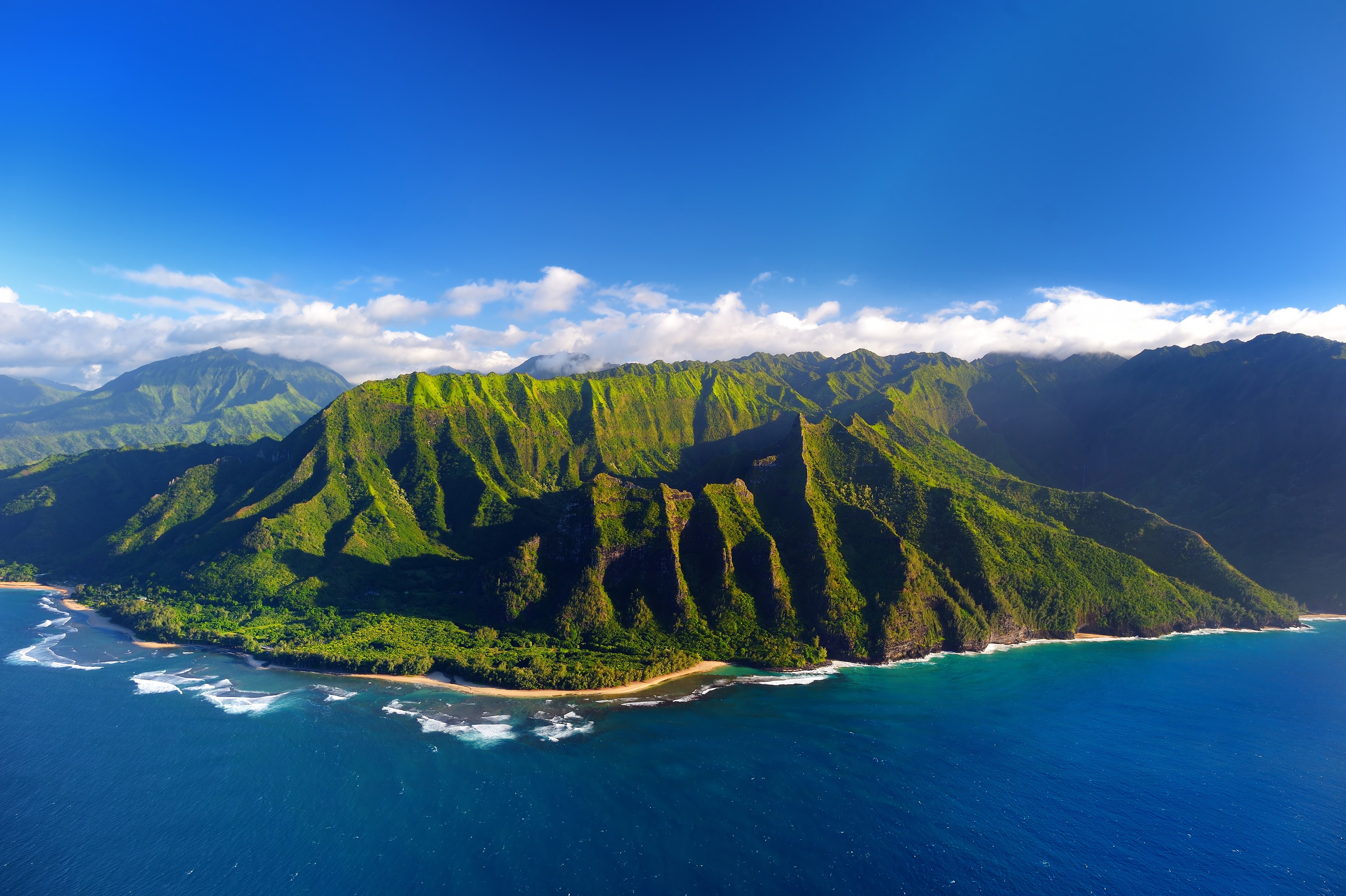 Luftfoto af de frodige, grønne klipper ved Na Pali-kysten på Kauai Island, Hawaii, med Stillehavet i forgrunden under en klar blå himmel