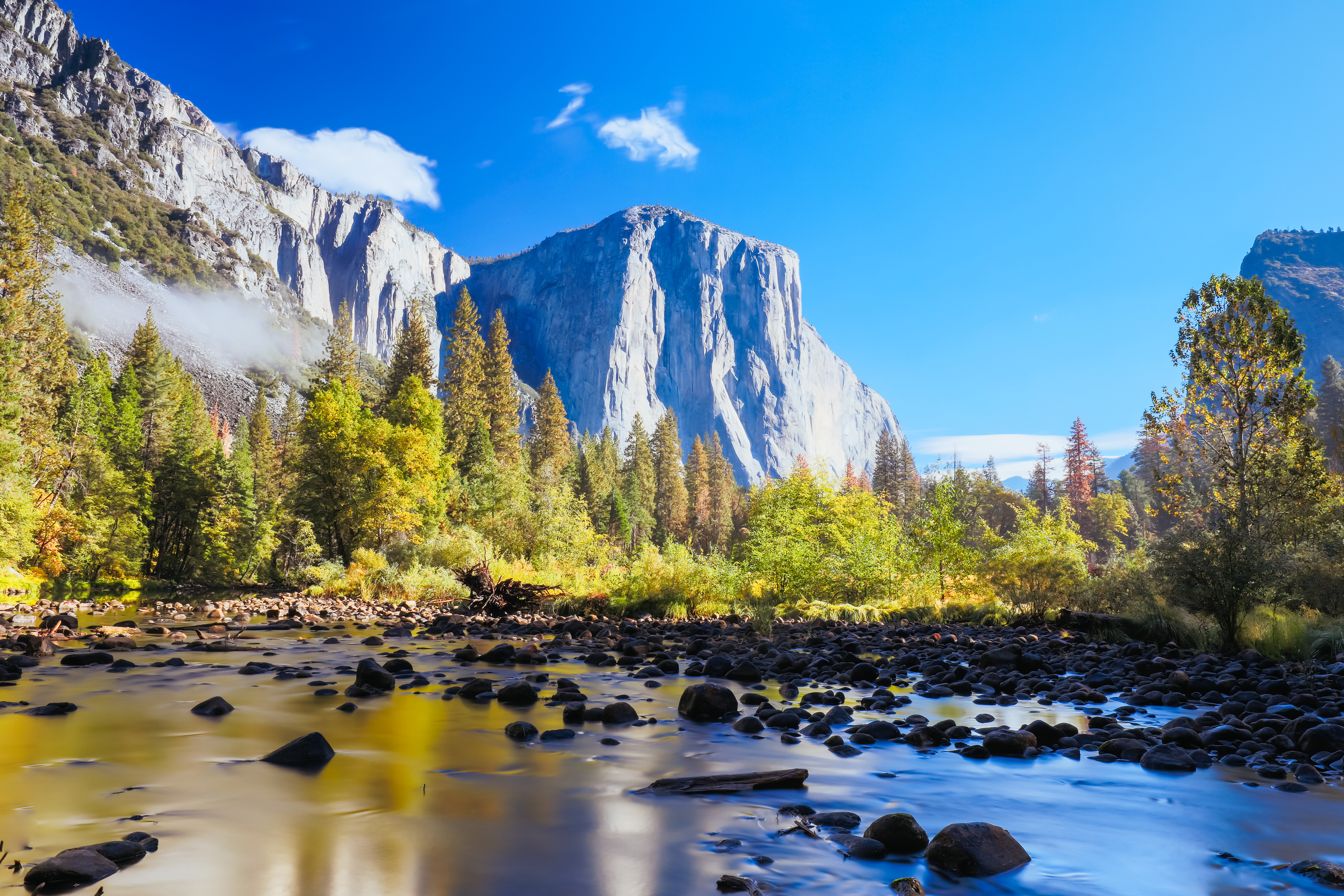 Udsigt over El Capitan i Yosemite National Park med en klar blå himmel, frodige grønne træer og en rolig flod i forgrunden