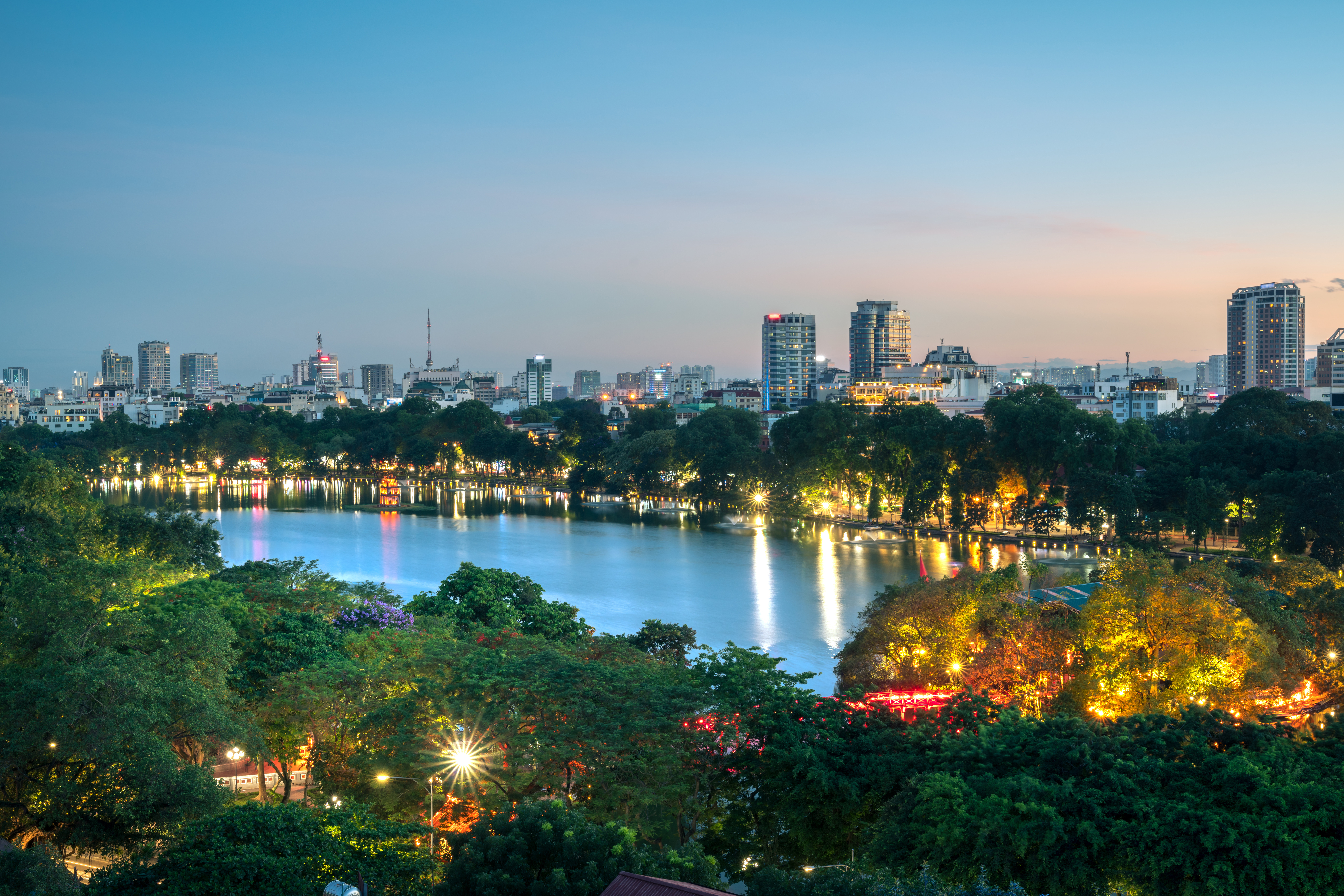 Skyline of Hanoi, Vietnam at dusk with Hoan Kiem Lake in the foreground, surrounded by illuminated urban buildings and vibrant greenery