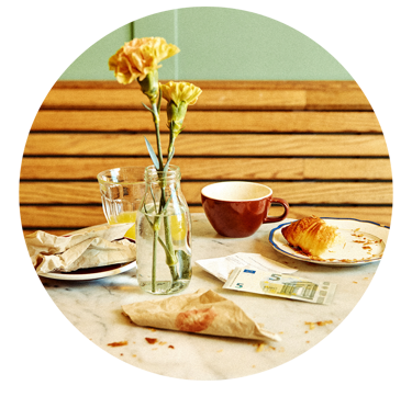Morgenbord med blomster i en vase, en kop, croissant på en tallerken og en fem-euroseddel.