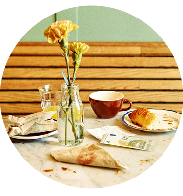 Blomster i en vase ved siden af en kaffekop, croissant og euroseddel på caféborde.