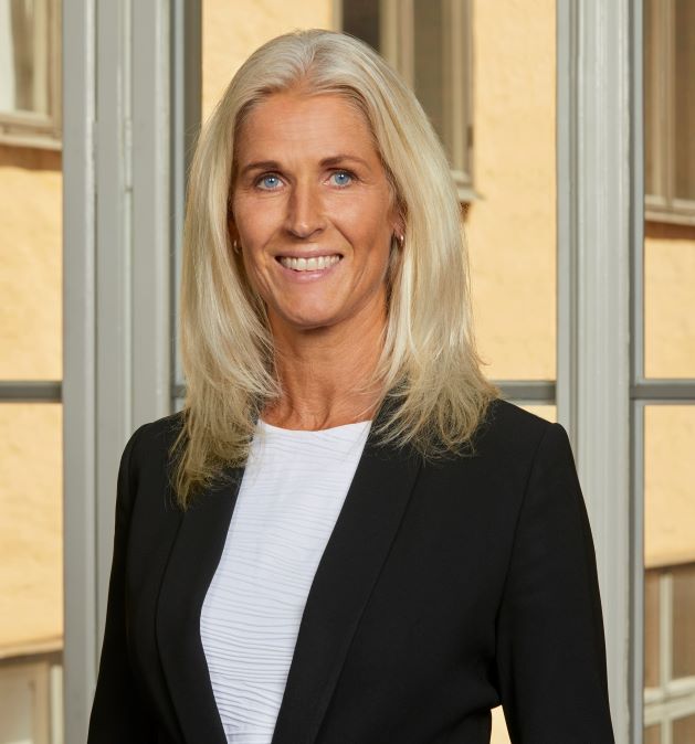 En kvinde med blondt hår i en sort jakke står foran et vindue.