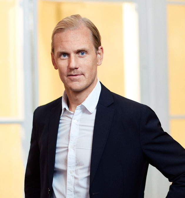 Manden i sort jakke og hvid skjorte står foran lyse vinduer.