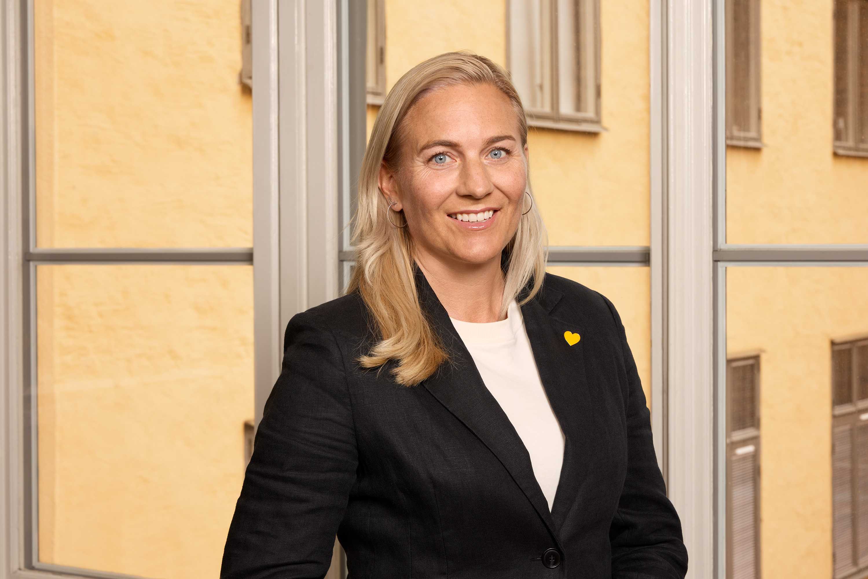 Kvinde i jakkesæt med gul nål står i et rum med store vinduer og gule vægge.