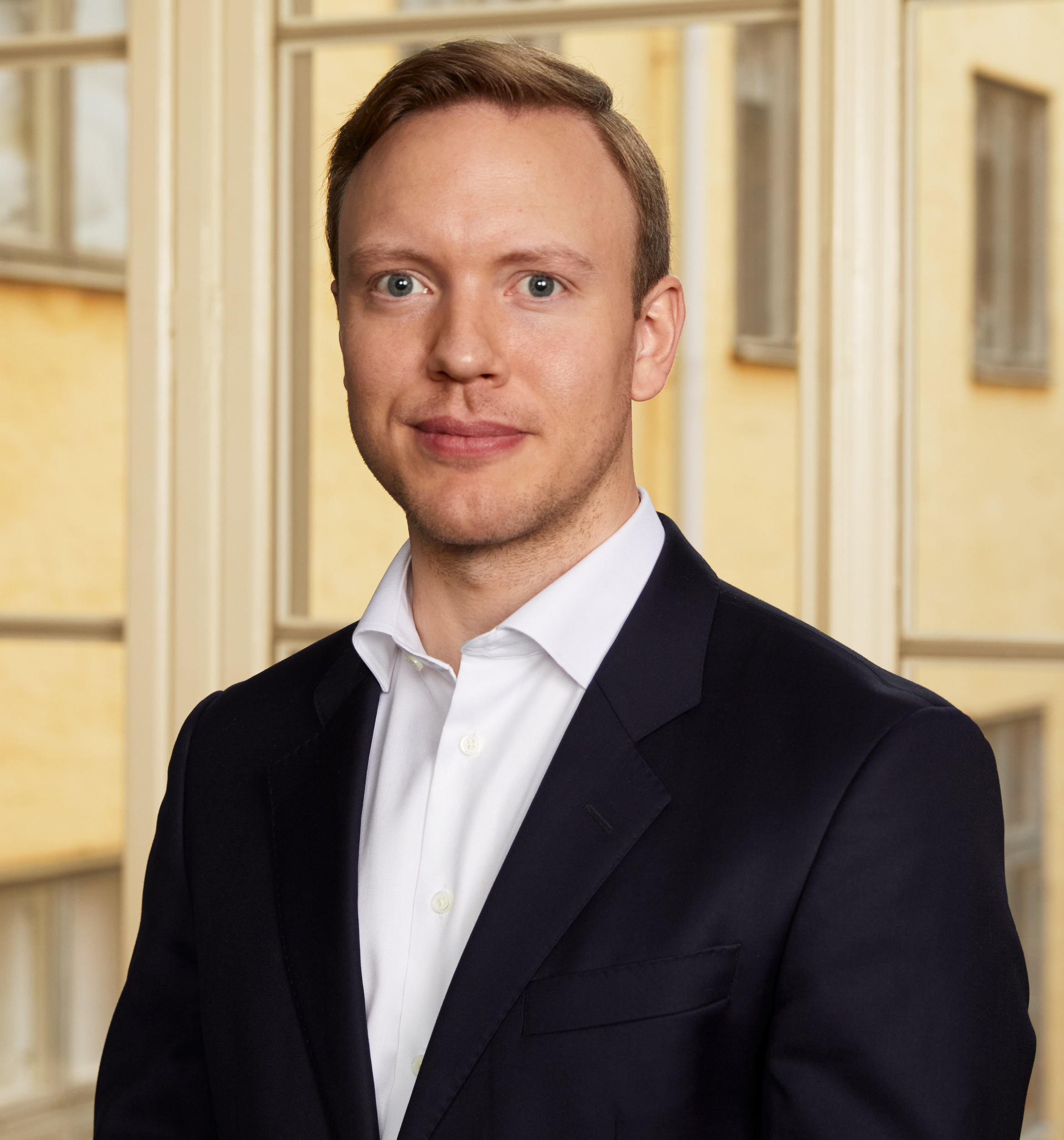 Person i jakkesæt med kort blondt hår foran en sløret baggrund af bygninger.