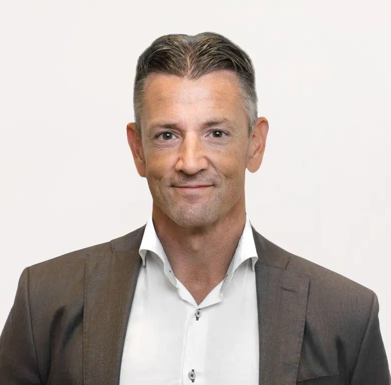 Man in a brown suit with a white shirt, smiling at the camera against a plain background.