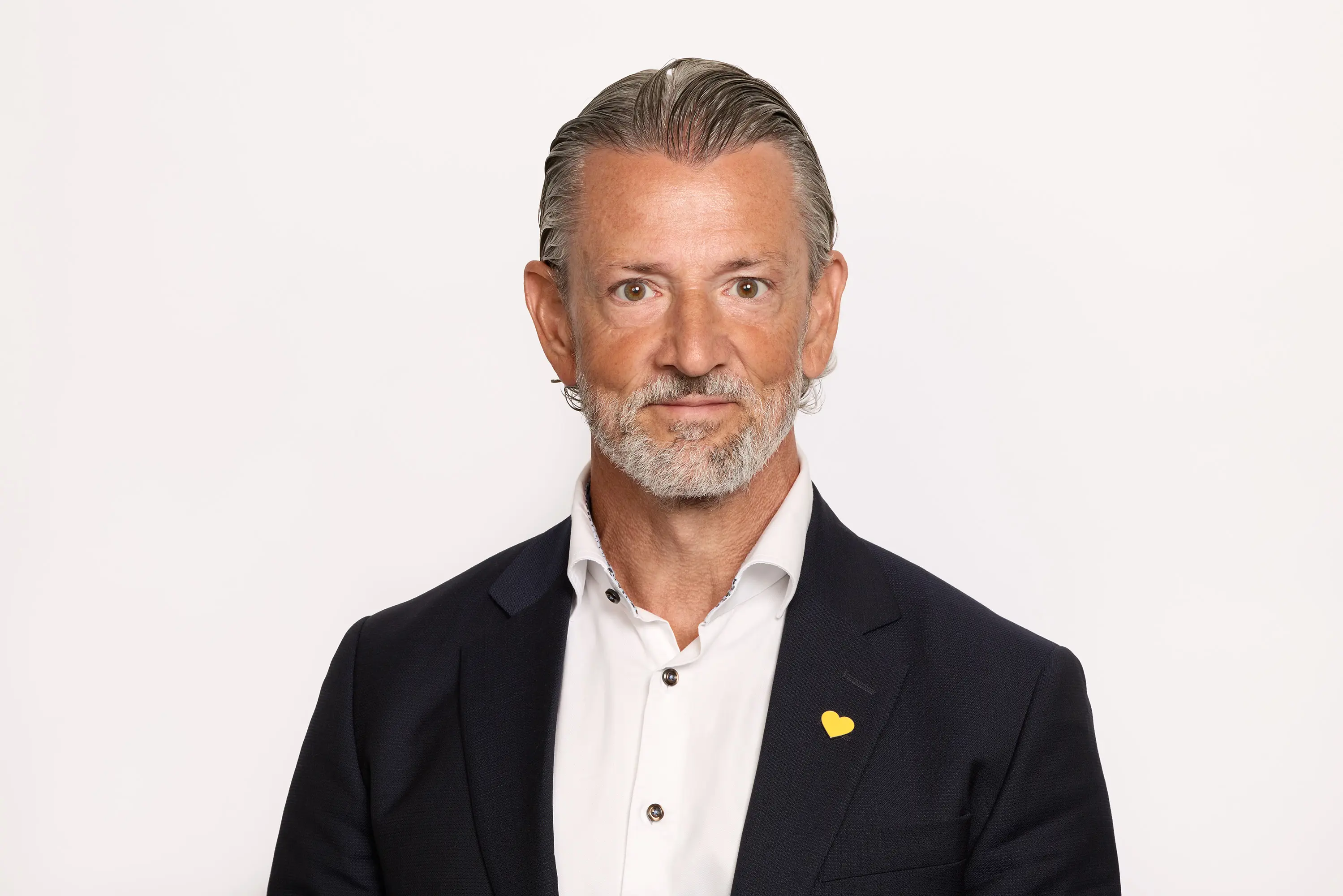 En mand i jakkesæt med skæg og kort gråt hår, iført en hvid skjorte og en gul nål formet som et hjerte på jakken.