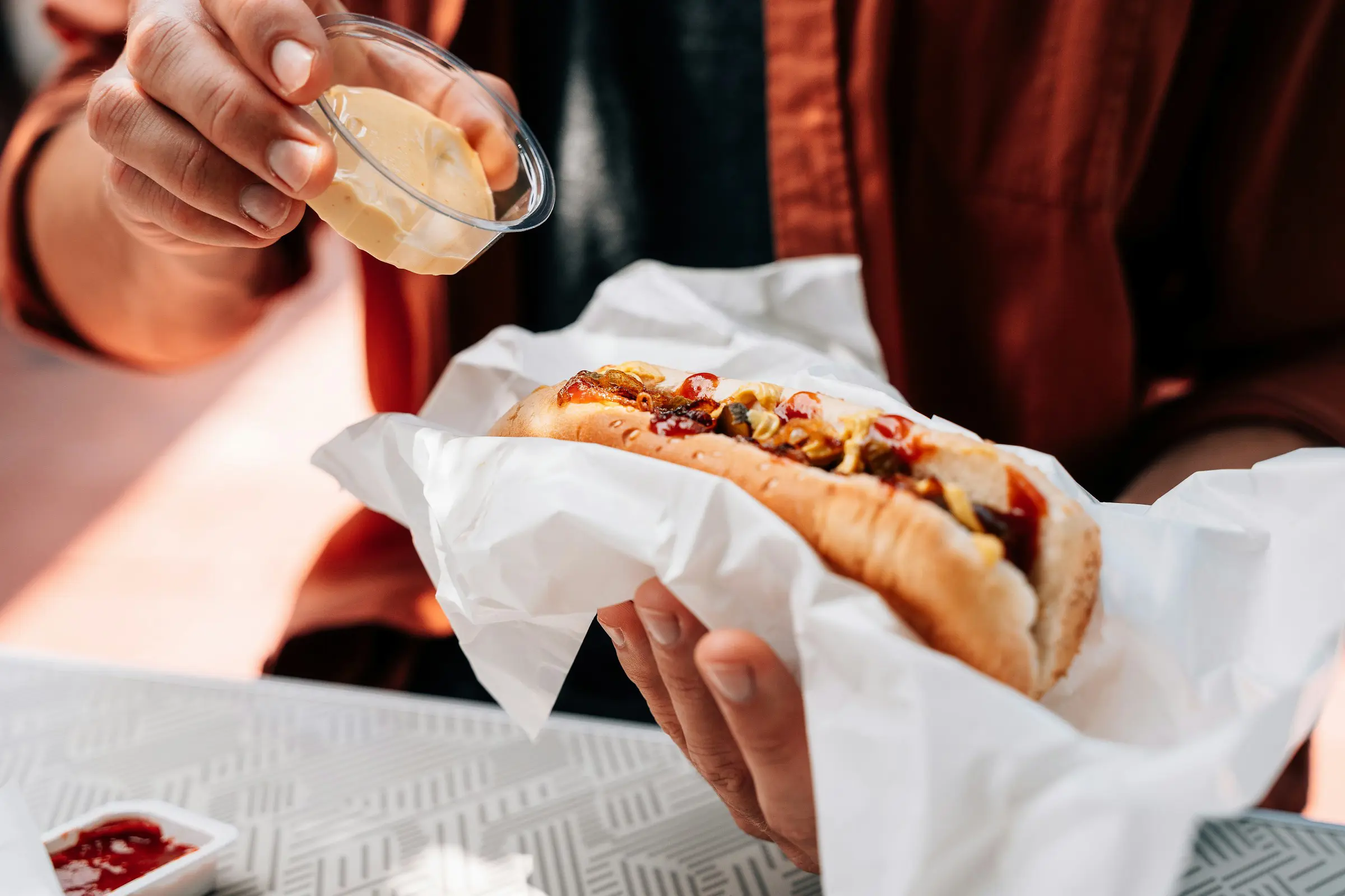 Person tilføjer sennep til hotdog pakket i papir på cafébord.