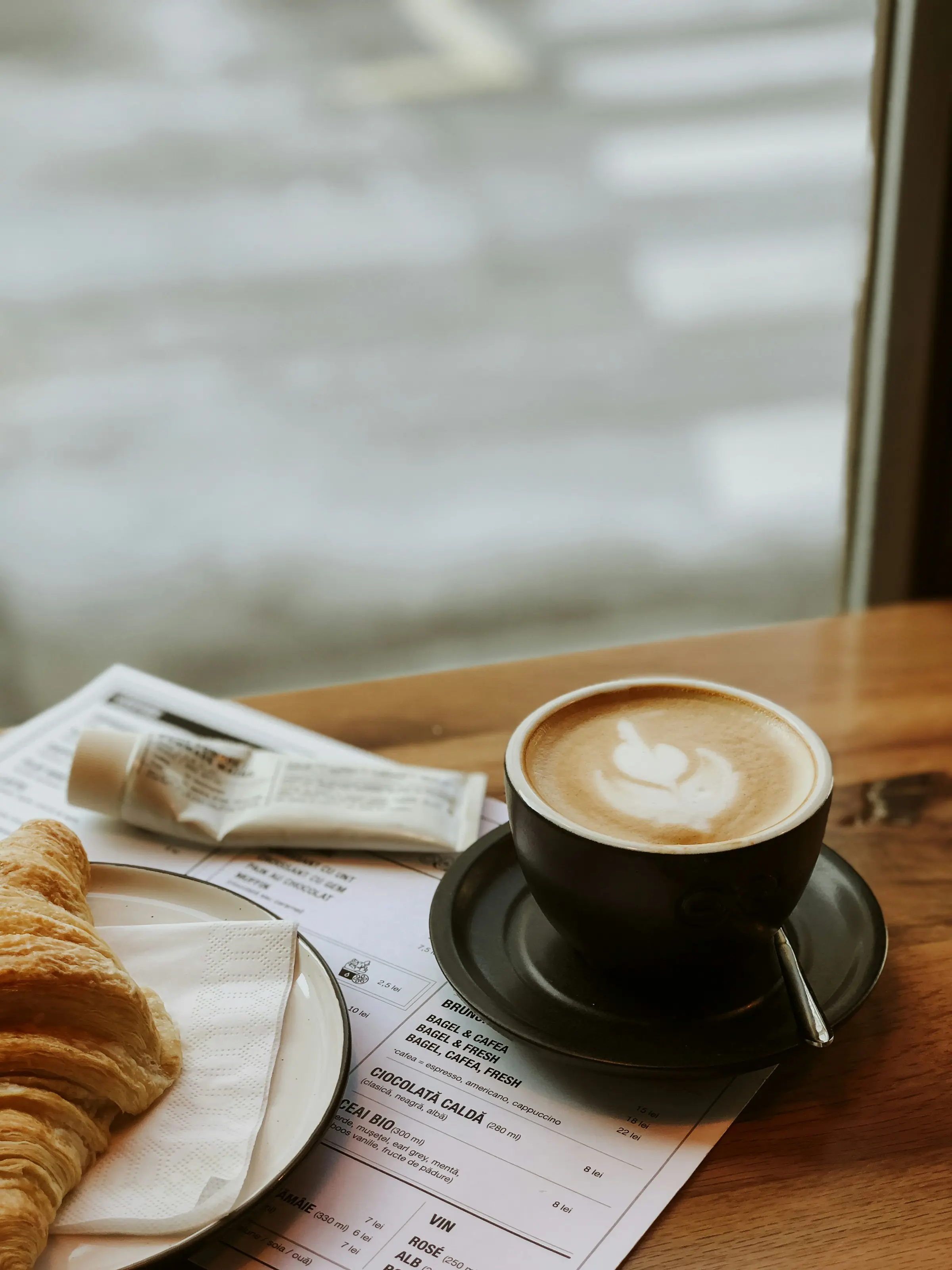 Cappuccino og croissant på træbord med menu i baggrunden. Perfekt til morgenmad eller kaffe på en café.