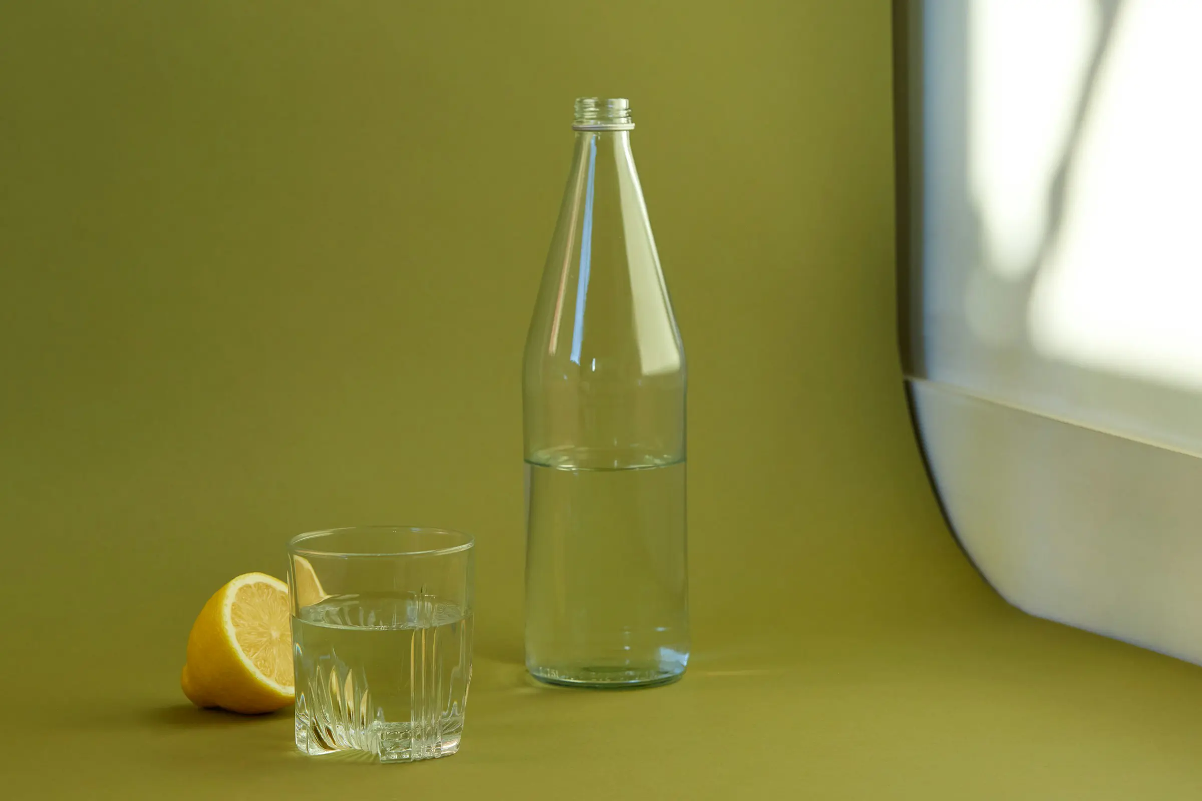 Glasflaske og glas vand ved siden af en halveret citron på et grønt bord.