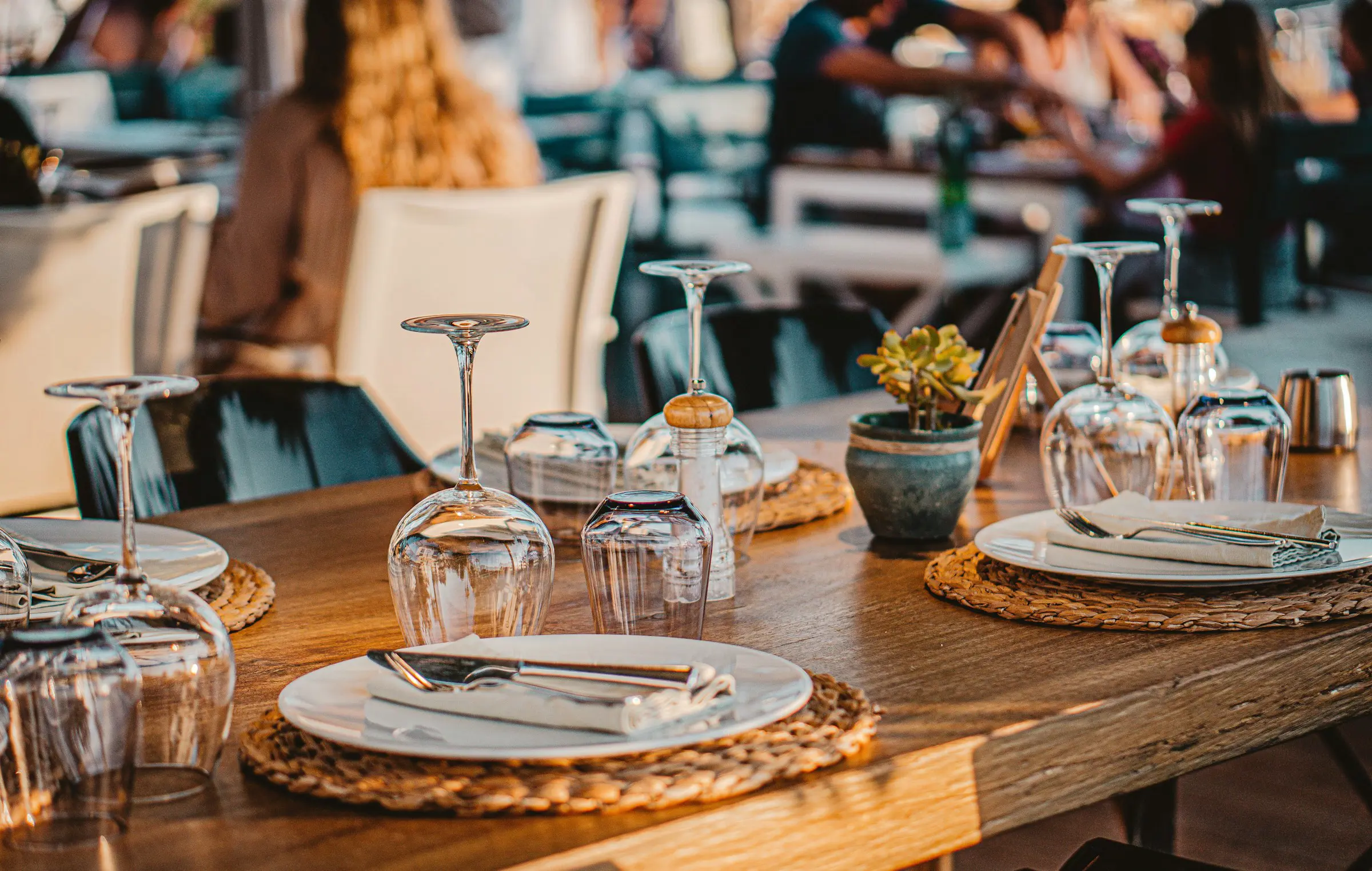 Smukt dækket bord i en restaurant med glas, tallerkener og bestik på flettede dækkeservietter, elegant atmosfære.