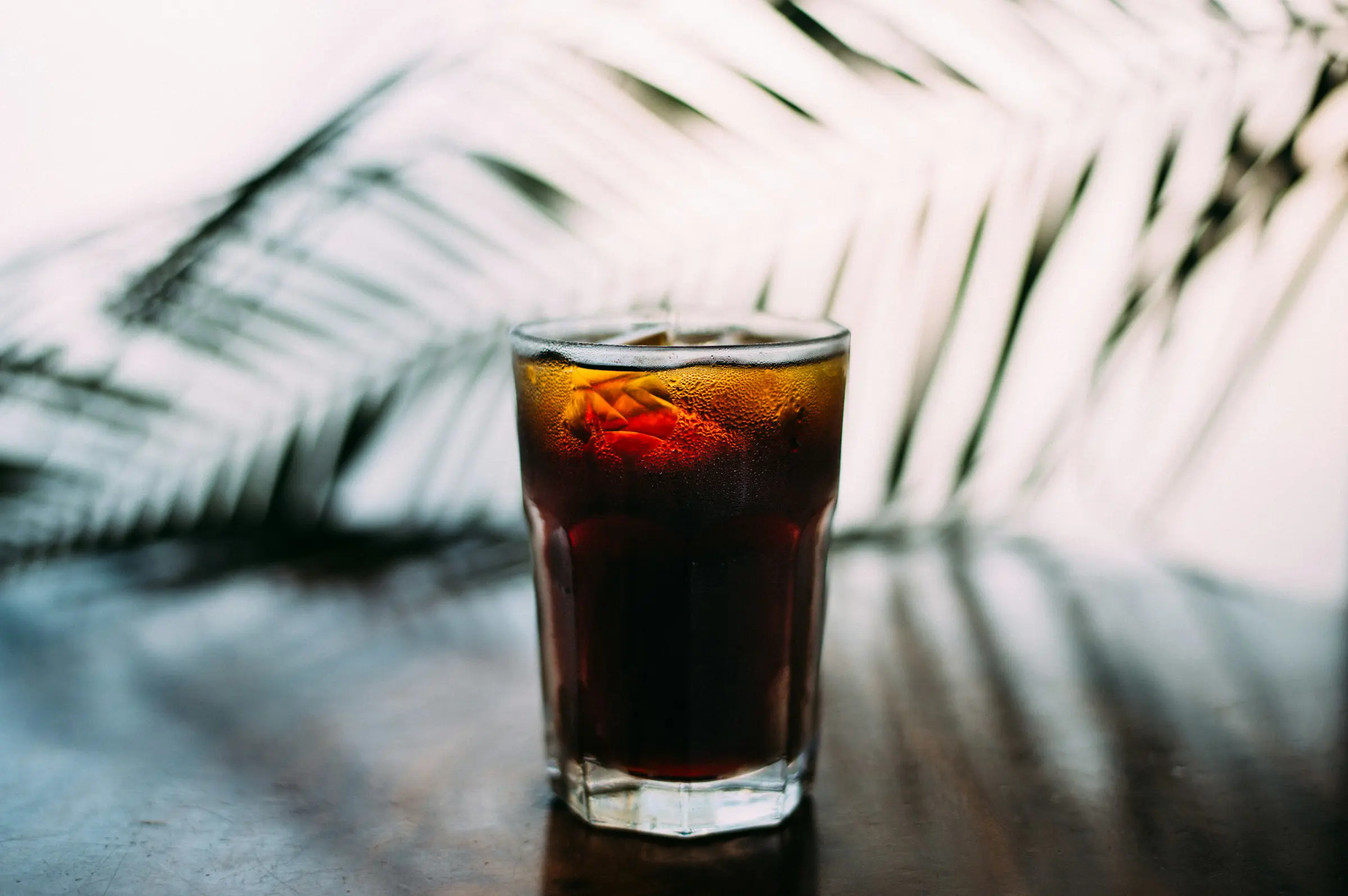 Glas med kold sodavand på bord med palmeblade i baggrunden.