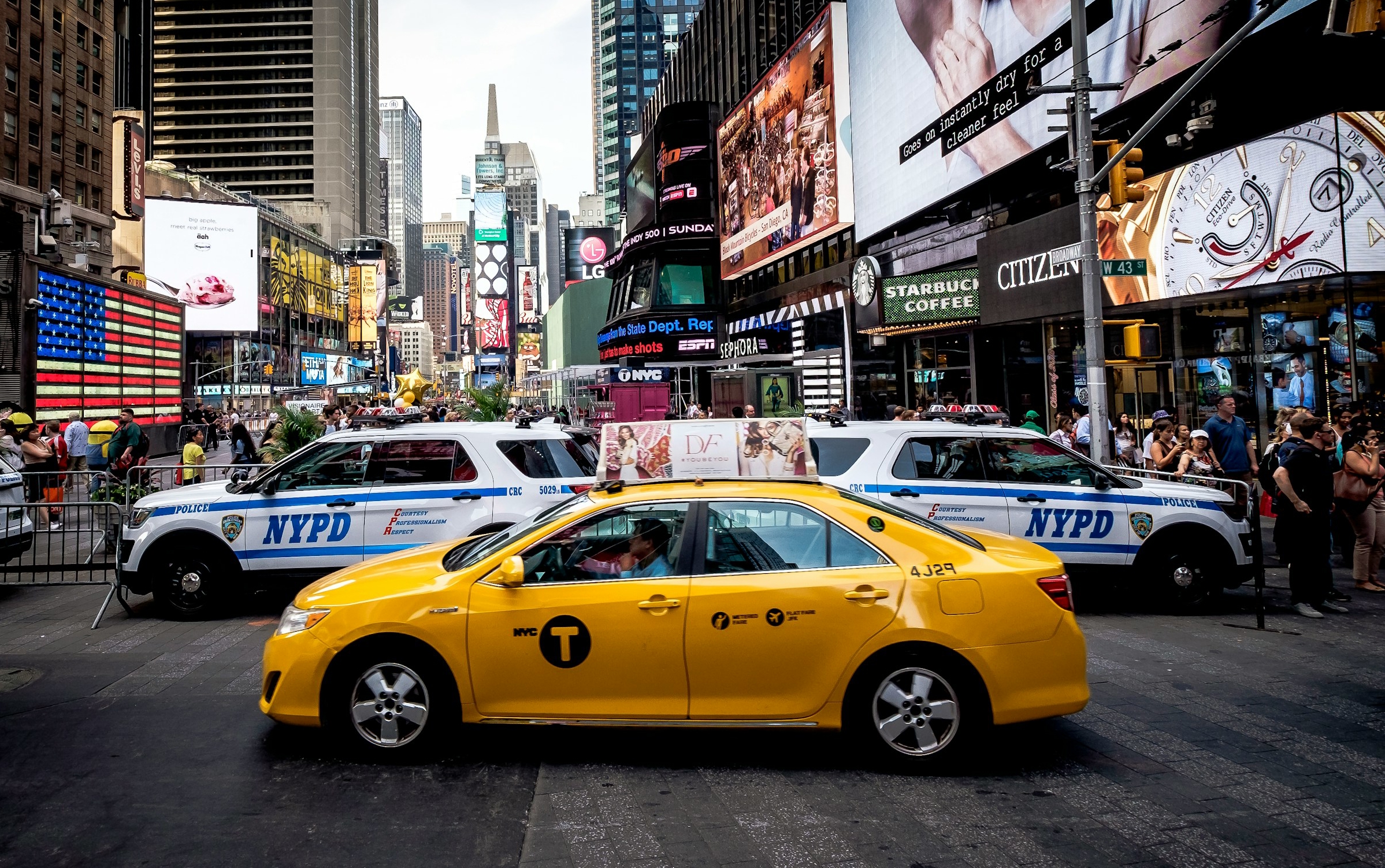 Gul taxa kører gennem Times Square i New York City med to NYPD-politibiler i baggrunden, omgivet af lysreklamer og butikker.