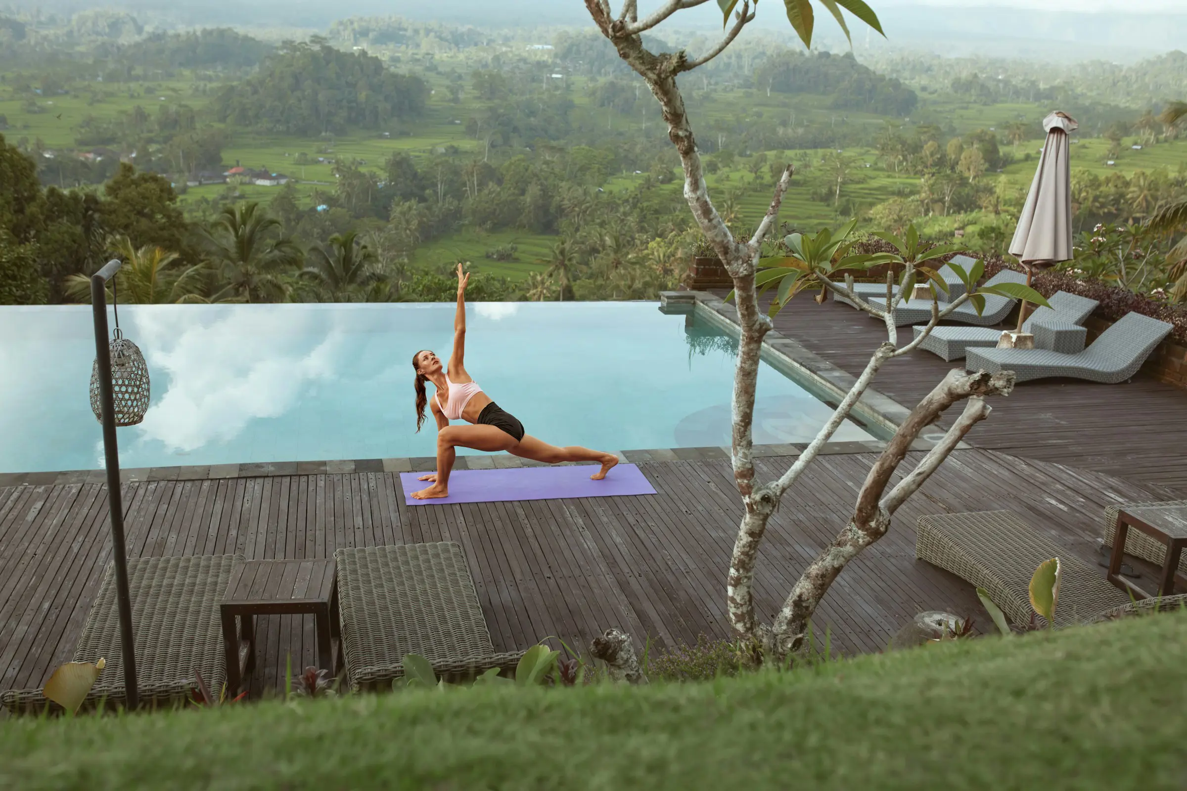 Woman practicing yoga on a mat by an infinity pool with a scenic view of lush green hills and farmland.